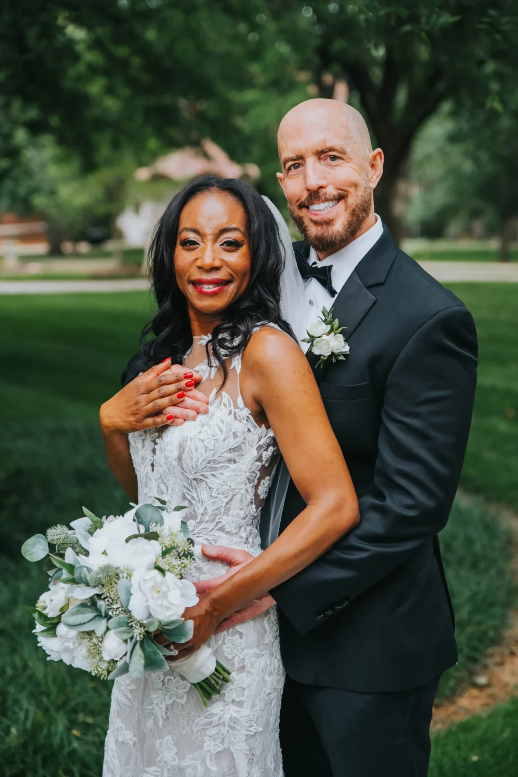 Elopement Wedding A couple on their wedding day. The bride, in a sleeveless lace gown, holds a bouquet of white flowers and stands in front of the groom, a man in a black tuxedo. They are outdoors in a green, park-like setting. Both are smiling and embracing, with trees and a blurred background behind them. Elopements Inc