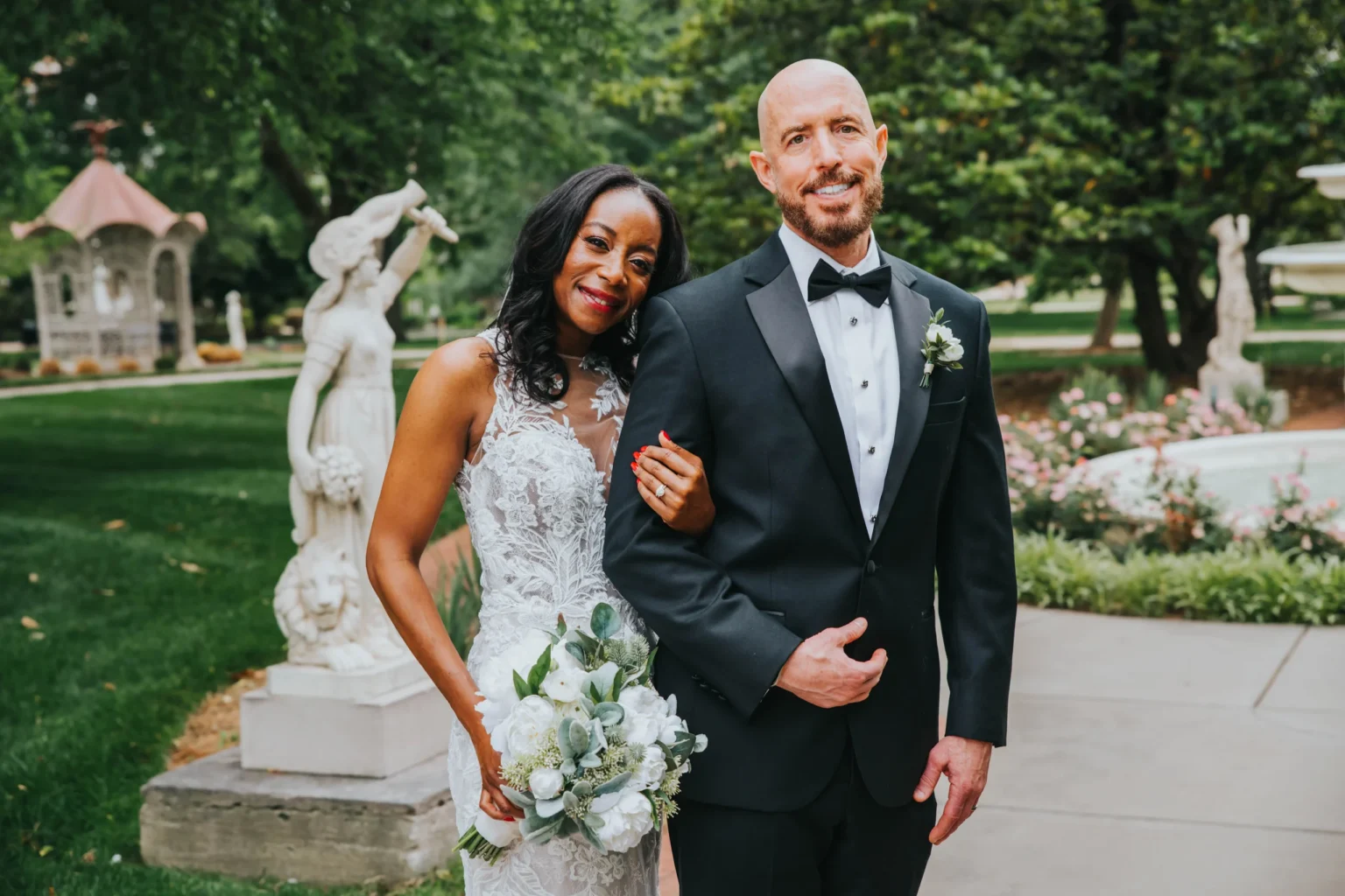 Elopement Wedding A newlywed couple posing outdoors in front of a garden. The bride, in a white lace gown, holds a bouquet of white flowers and smiles warmly, standing with her arm around the groom's. The groom, wearing a black tuxedo with a bow tie and boutonniere, looks content. Statues and greenery are in the background. Elopements Inc