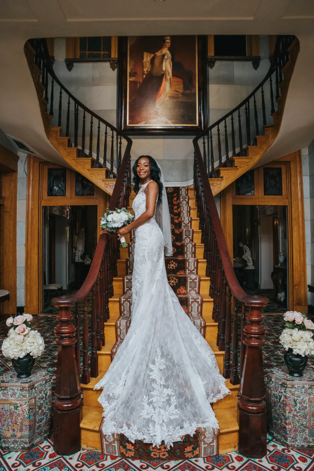 Elopement Wedding A bride in a detailed white lace gown and veil stands on a grand wooden staircase holding a bouquet of flowers. The staircase features ornate railings and leads to a landing with a large portrait hanging above. Flower arrangements are seen at the base of the staircase. Elopements Inc