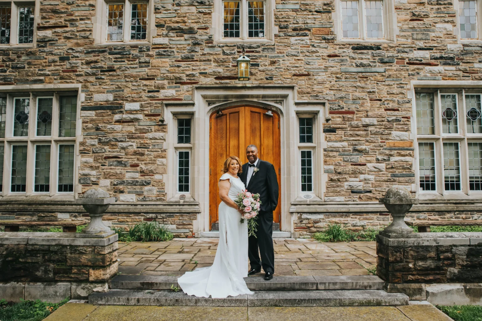 Elopement Wedding A bride and groom stand in front of a historic stone building with a wooden door and arched windows. The bride is in a white gown holding a bouquet of pink and white flowers, while the groom is in a black tuxedo. Both are smiling and posed in the center of the image on a stone pathway. Elopements Inc