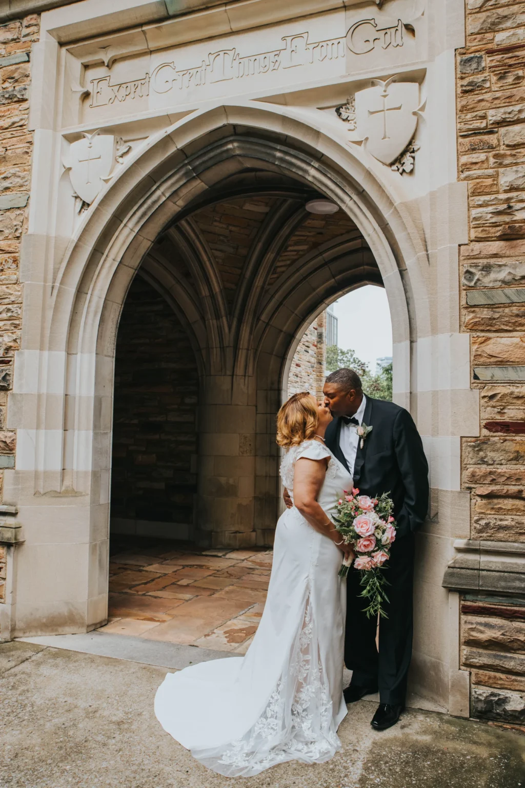 Elopement Wedding A newlywed couple shares a kiss under a stone archway. The bride wears a white dress with lace details and holds a bouquet of pink and white flowers. The groom is in a dark suit with a boutonnière. Above them, an engraved phrase reads "Expect Great Things From God. Elopements Inc