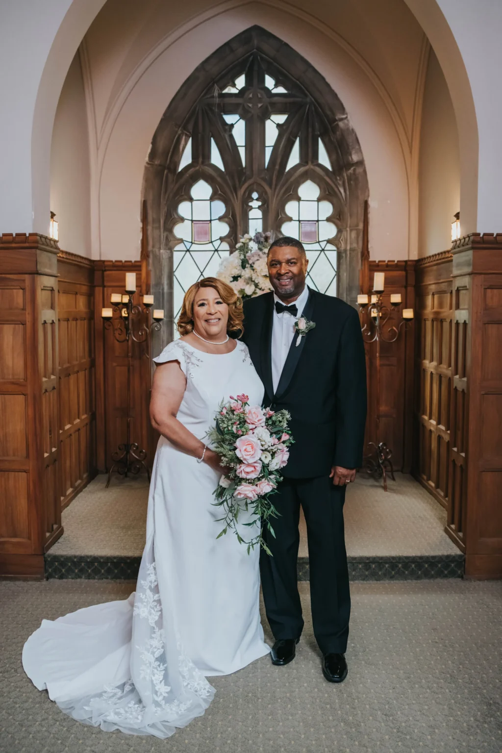 Elopement Wedding A smiling couple stands together in a wooden-paneled chapel with tall arched windows. The bride wears a white gown and holds a cascading bouquet of pink and white flowers. The groom is dressed in a black tuxedo with a white shirt and a boutonniere. They stand close, radiating happiness. Elopements Inc