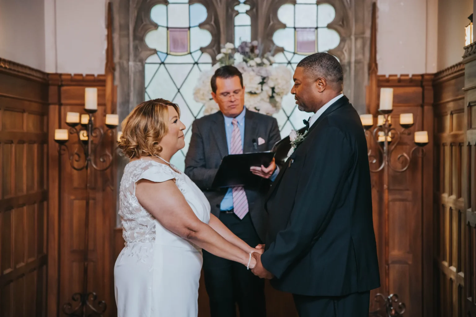 Elopement Wedding A couple stands holding hands during their wedding ceremony in a wood-paneled room. The bride, in a white dress, faces the groom, who is in a black suit. They are gazing at each other while an officiant, standing behind them, has an open book. A stained glass window is visible in the background. Elopements Inc