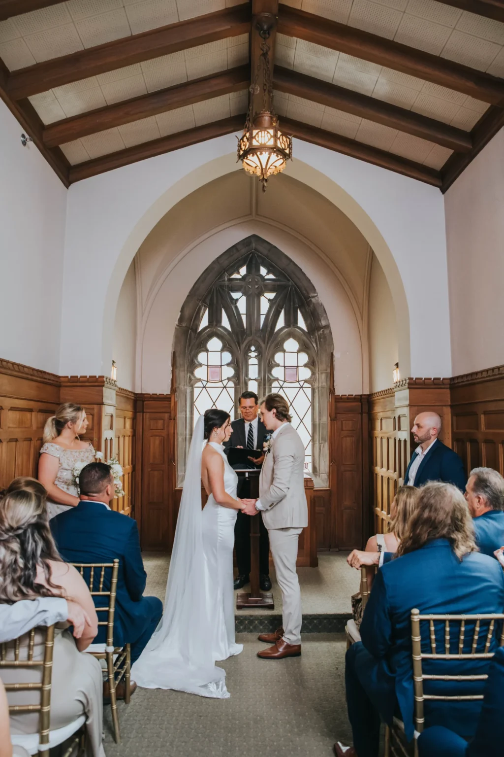 Elopement Wedding A couple stands at the altar during their wedding ceremony in a small chapel. The bride is wearing a white dress and veil, holding hands with the groom in a light-colored suit. The officiant stands behind them. Guests are seated on either side, watching the couple. The chapel features wooden paneling and arched windows. Elopements Inc