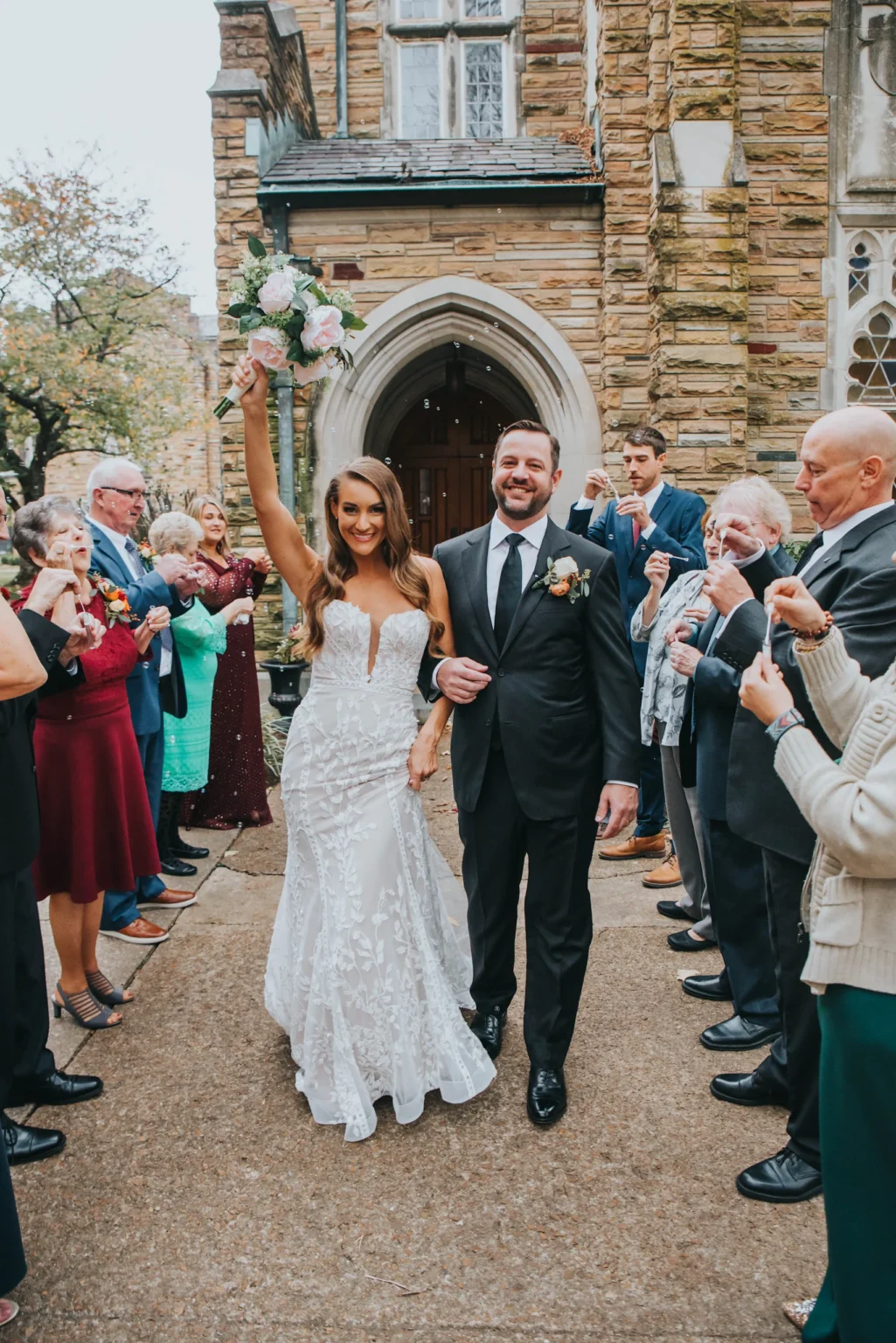 Elopement Wedding A newly married couple walks out of a stone church, smiling and holding hands. The bride, wearing a white lace dress, holds a bouquet high in the air. The groom, in a black suit and tie, beams beside her. Guests line the path, cheering and taking photos, some blowing bubbles. Elopements Inc