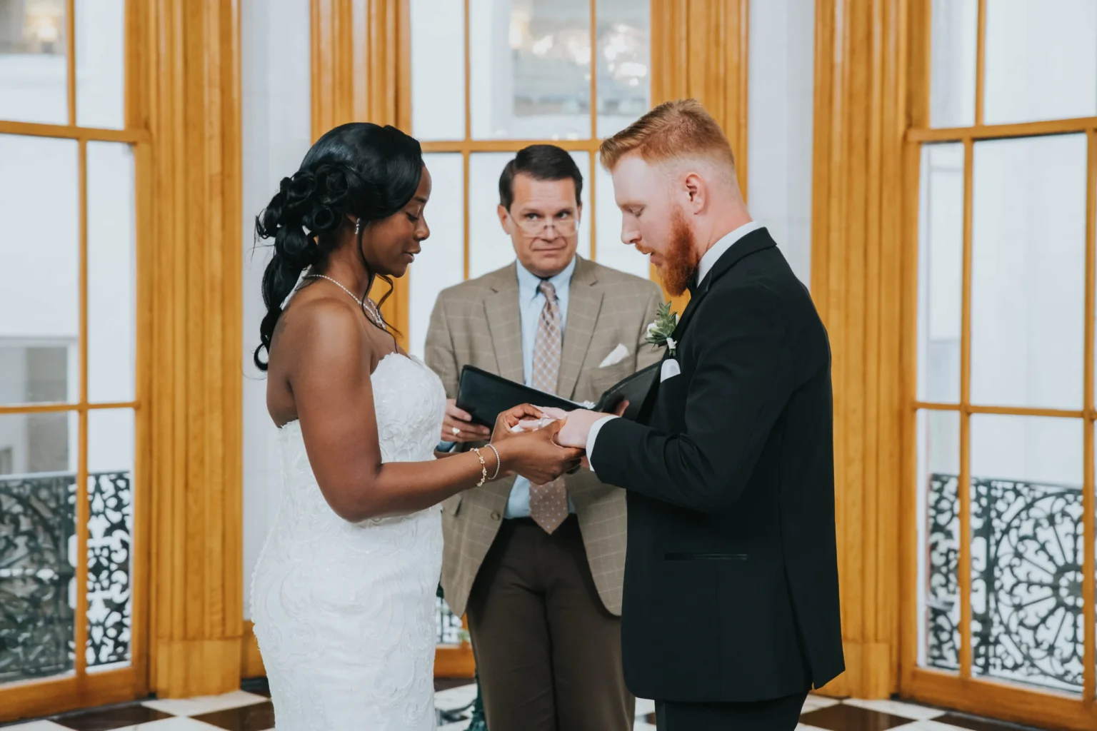 Elopement Wedding A bride and groom stand facing each other in a room with large windows and wooden frames. The bride, in a white dress, holds a ring while the groom, in a black suit, looks at it intently. Behind them, a man in a beige suit and blue tie officiates the ceremony. Elopements Inc