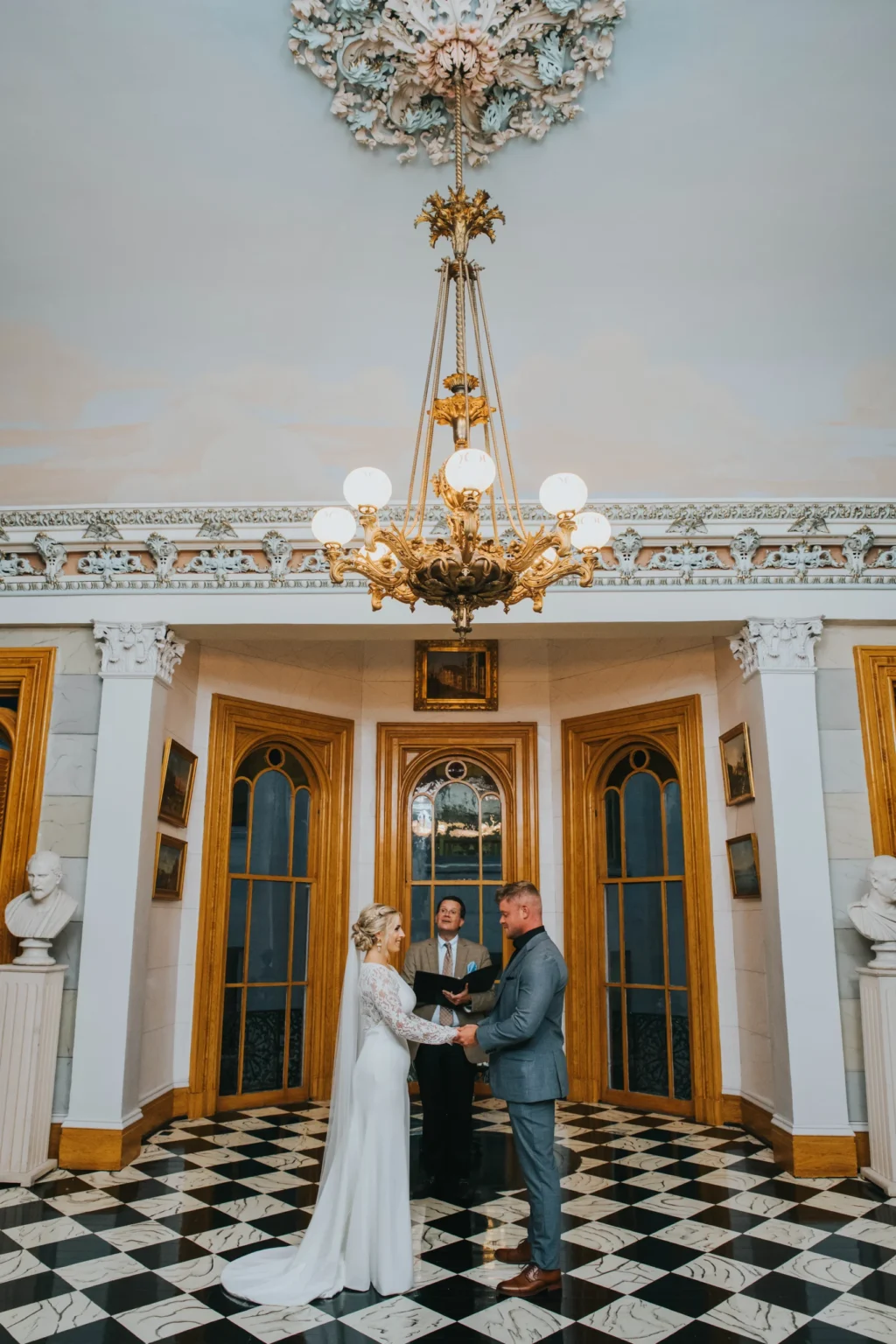 Elopement Wedding A couple stands in an ornate room under a grand chandelier, facing each other and holding hands, with a person officiating their wedding in the background. The room features marble flooring, gold trim, tall windows, and busts on pedestals. The bride wears a long white dress and the groom a gray suit. Elopements Inc