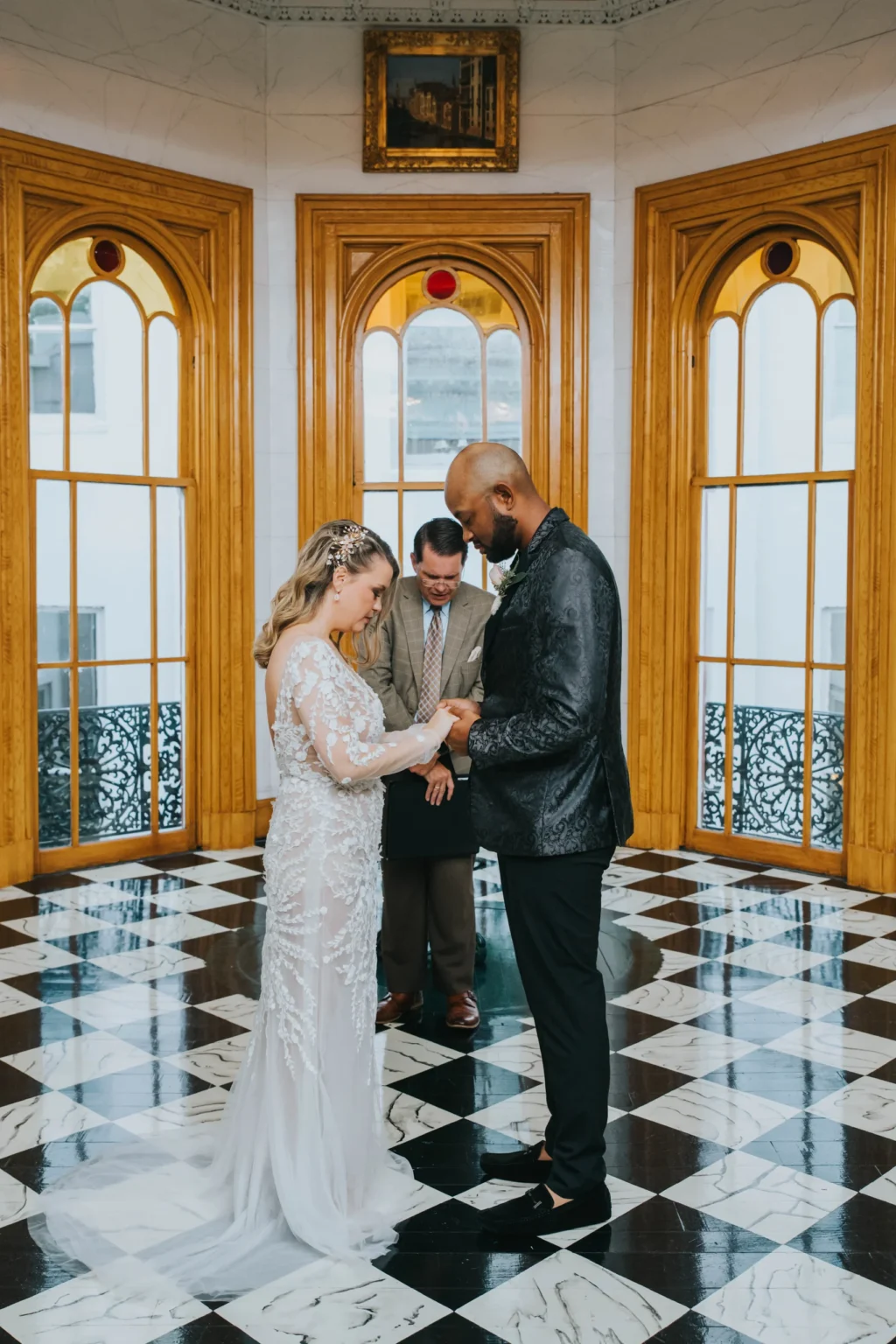 Elopement Wedding A couple stands facing each other holding hands in a beautifully ornate room with large wooden-framed windows and a black-and-white checkered marble floor. The bride is in a white dress with lace details, and the groom wears a black suit. A man in a suit stands behind them, officiating. Elopements Inc