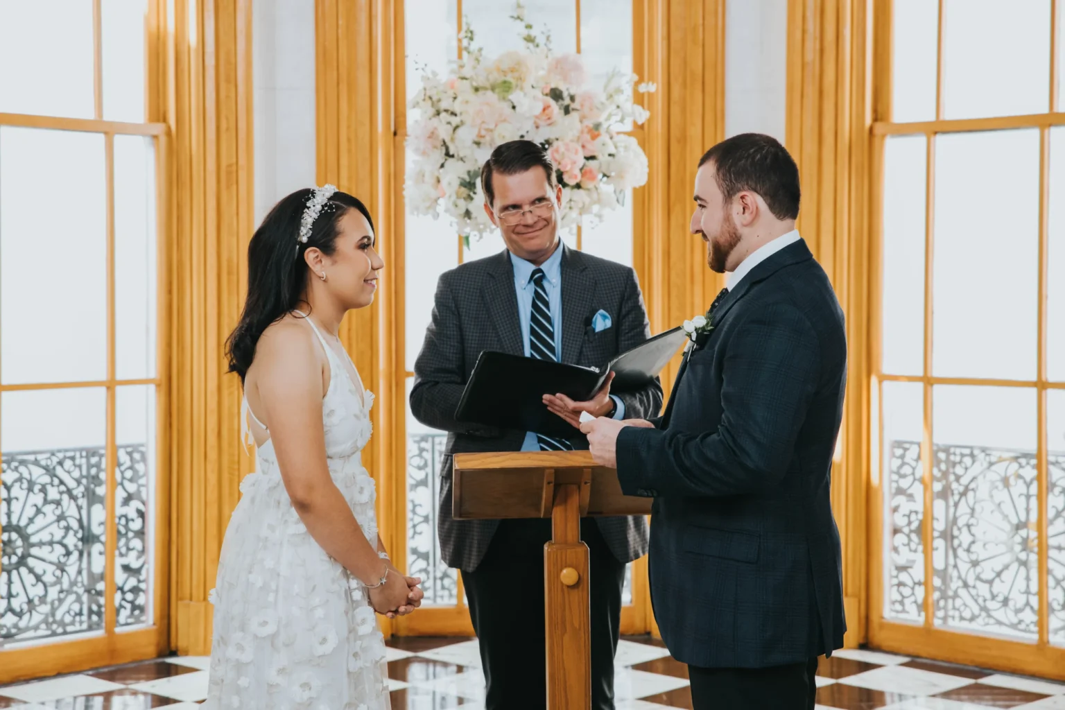 Elopement Wedding A couple is exchanging wedding vows in an elegant room with large windows and wooden frames. The bride wears a white floral dress and headband, and the groom is in a dark blue suit. An officiant stands between them, holding a book and smiling. Floral arrangements are in the background. Elopements Inc