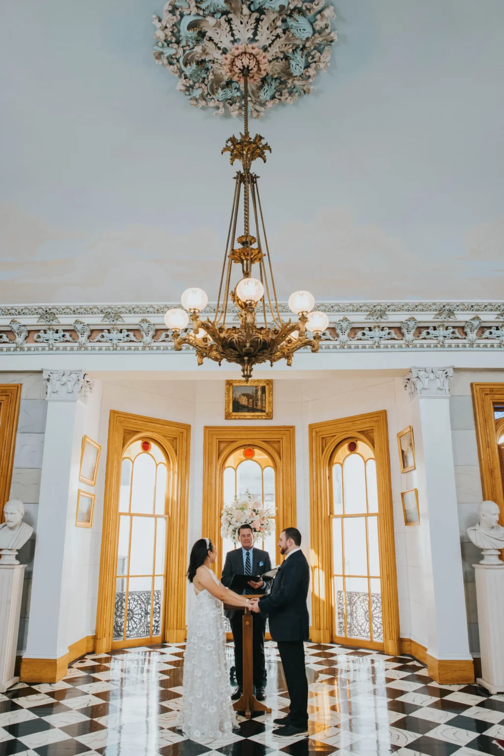 Elopement Wedding A couple stands facing each other during their wedding ceremony in an elegant room. An officiant stands before them holding a book. The setting features ornate arches, decorative moldings, and a grand chandelier hanging from a high ceiling. Marble busts are placed on pedestals along the walls. Elopements Inc