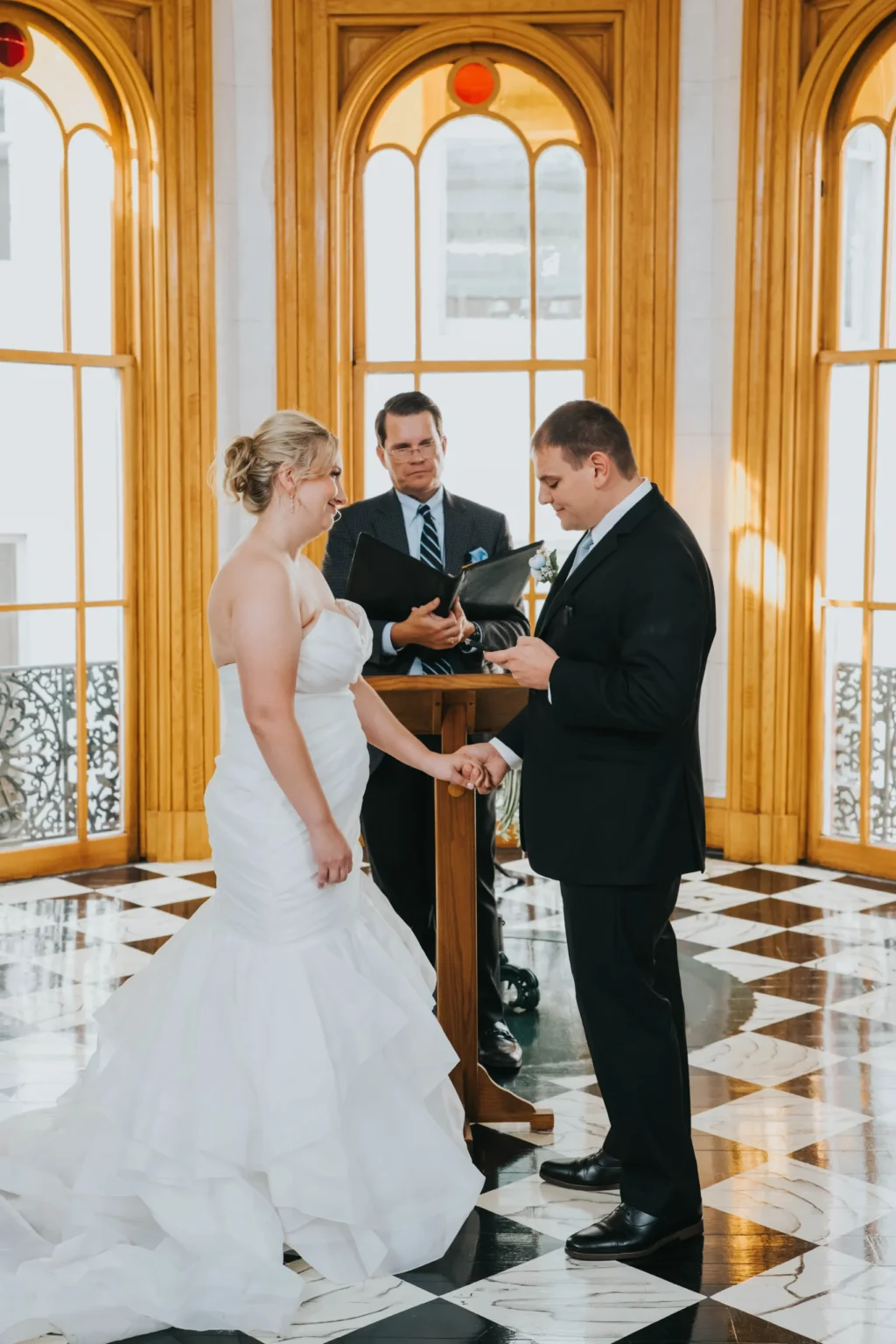 Elopement Wedding A couple, dressed in wedding attire, stands facing each other and holding hands in a sunlit room with large arched windows and wooden frames. The groom holds a ring. They are accompanied by an officiant standing behind a wooden lectern. The black-and-white checkered floor adds elegance to the scene. Elopements Inc