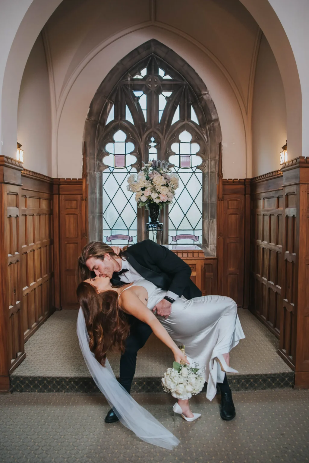 Elopement Wedding A bride in a flowing white dress and a groom in a black suit share a dramatic kiss in a wood-paneled room with arched ceilings. The groom dips the bride, who holds a bouquet and wears a veil. A large bouquet of flowers is displayed in front of a stained glass window behind them. Elopements Inc