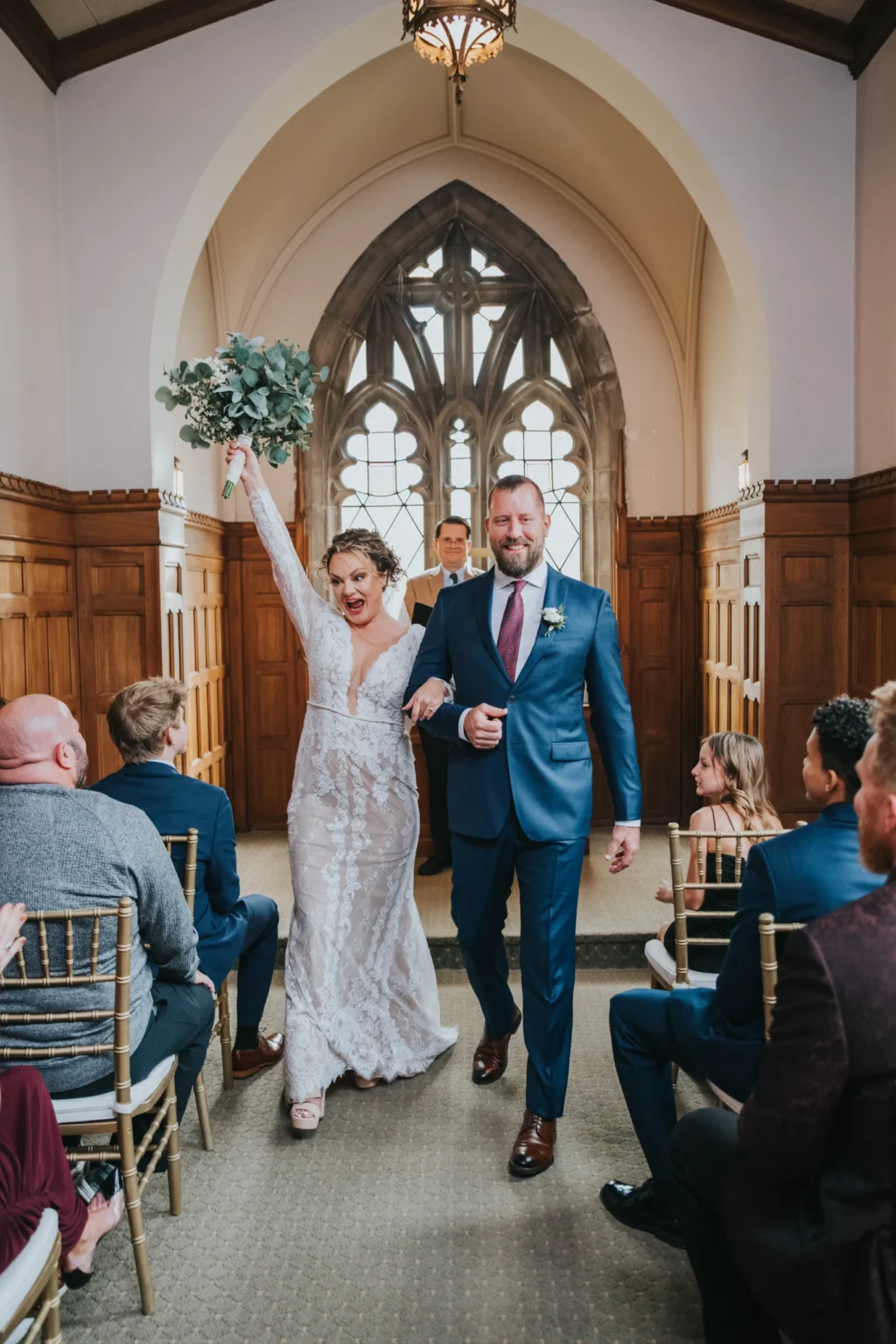 Elopement Wedding A bride in a long-sleeved white wedding dress holds a bouquet aloft while walking down the aisle with the groom, who is wearing a blue suit and tie. They are smiling, followed by a man in a suit. Guests, seated on both sides in wooden chairs, look on inside a hall with arched windows and wooden paneling. Elopements Inc