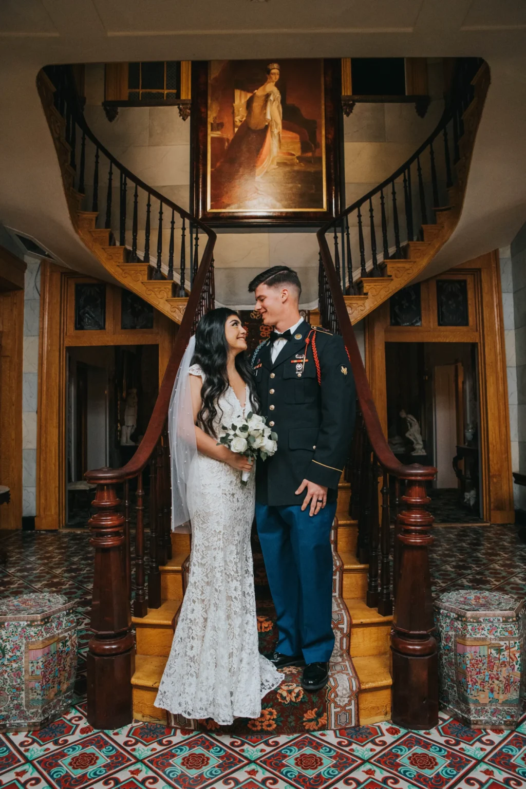 Elopement Wedding A bride in a white dress and veil holds a bouquet while standing with a groom in military uniform. They are gazing at each other in front of a grand wooden staircase with intricate railings. An elegant portrait painting hangs above. The floor is tiled with an ornate pattern. Elopements Inc