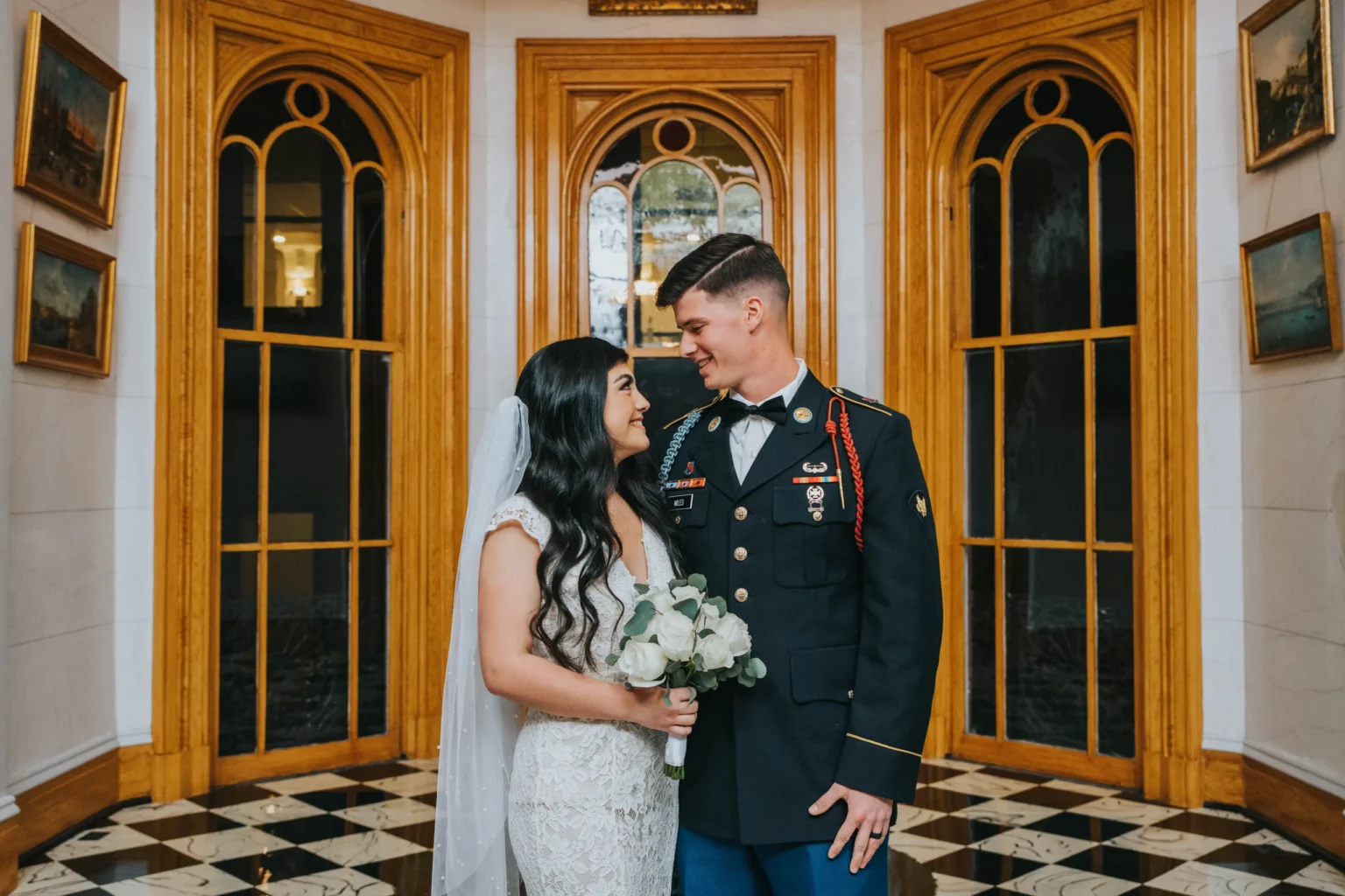 Elopement Wedding A bride in a lace wedding dress and veil stands next to a groom in a decorated military uniform. They are facing each other, smiling, holding a bouquet of roses. They are in a room with black and white checkered flooring, ornate wooden door frames, and several paintings on the walls. Elopements Inc