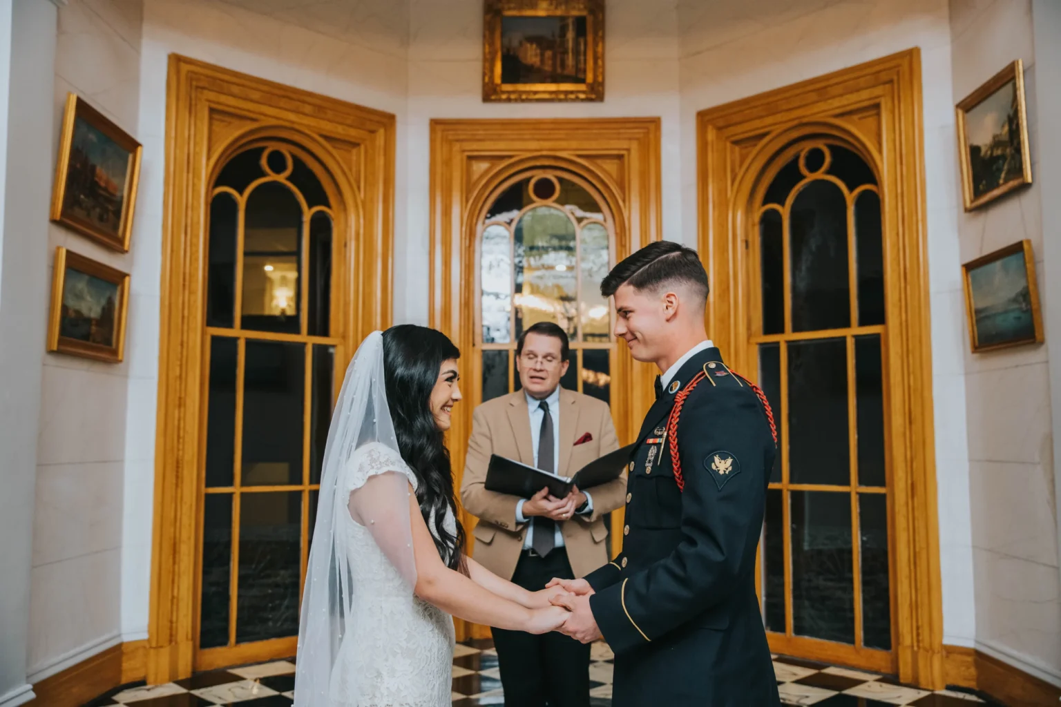 Elopement Wedding A bride and groom hold hands facing each other during their wedding ceremony. The groom is dressed in a formal military uniform, and the bride wears a white dress with a lace veil. An officiant stands behind them, holding a book. They are in an elegant room with arched wooden doors and paintings. Elopements Inc