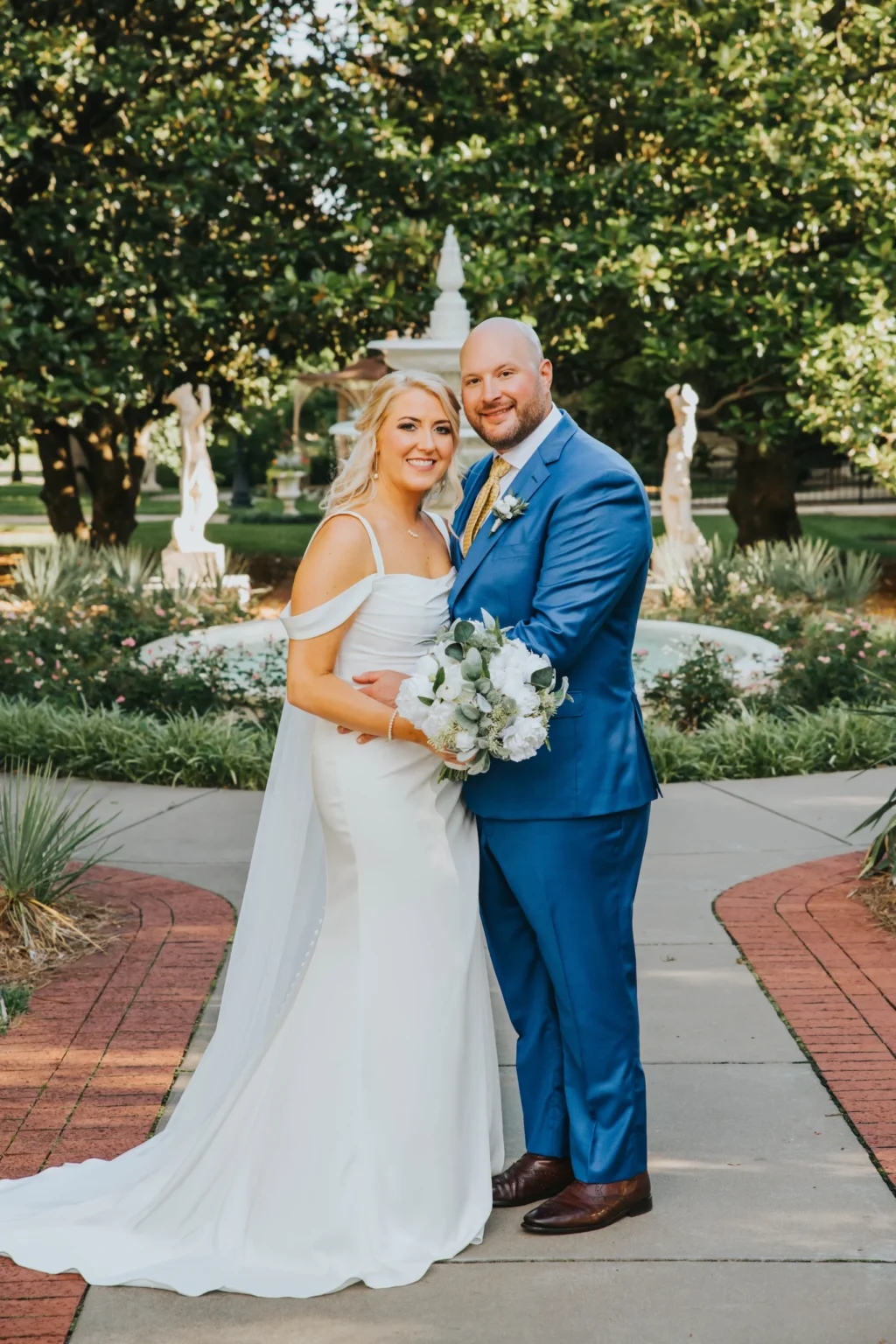 Elopement Wedding A bride and groom are posing outdoors on a sunny day, standing on a paved path with greenery and a white statue in the background. The bride is wearing a white off-shoulder wedding dress and holding a bouquet, while the groom is in a blue suit with a matching tie. They are smiling and looking at the camera. Elopements Inc