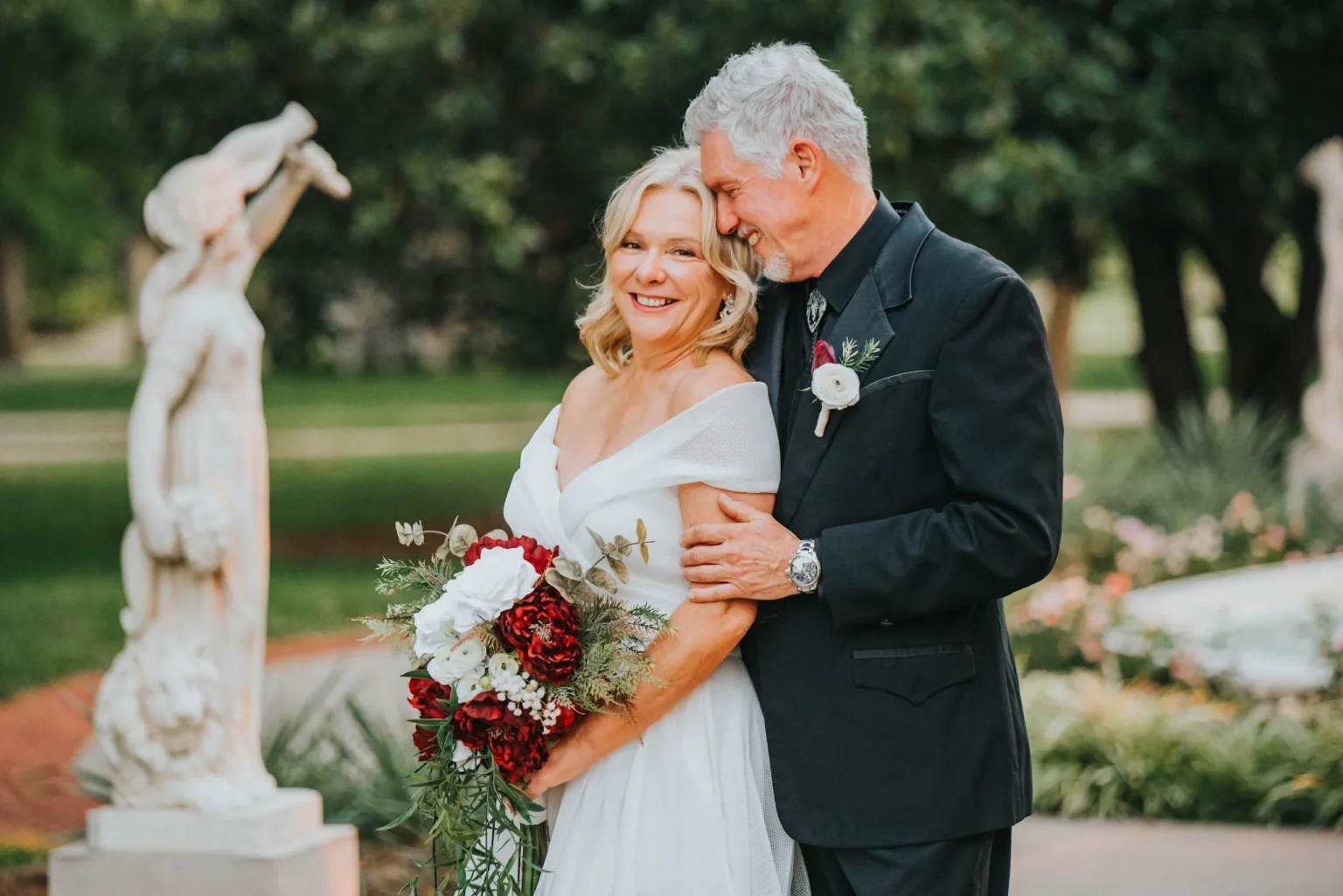 Elopement Wedding A smiling bride in an off-the-shoulder white gown holds a bouquet of red and white flowers, standing beside a groom in a dark suit. They are in an outdoor garden with statues in the background. The groom lovingly leans his head towards the bride’s, both appearing joyful and content. Elopements Inc