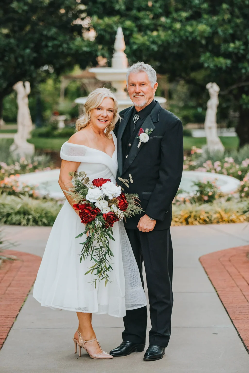 Elopement Wedding A smiling older couple stands outside in front of a fountain, embracing. The woman wears an off-the-shoulder white dress and holds a bouquet of red and white flowers. The man is dressed in a black suit with a white shirt and black tie. The background includes greenery and statues. Elopements Inc