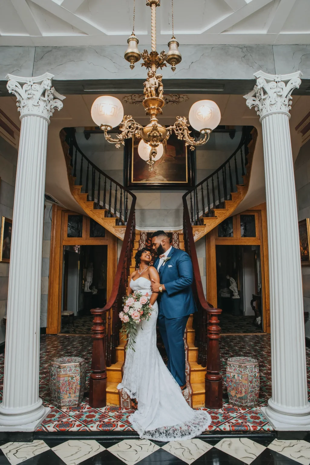 Elopement Wedding A bride in a white dress and groom in a blue suit stand on a grand staircase, holding hands and looking at each other warmly. The staircase is flanked by ornate columns and a vintage chandelier hangs above. The floor features a geometric black and white pattern. Elopements Inc