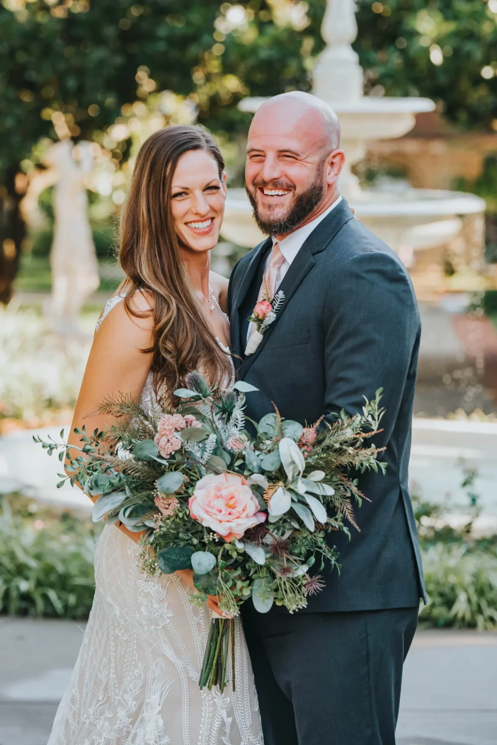 Elopement Wedding A smiling couple poses outdoors on their wedding day. The woman, wearing a white lace gown, holds a lush bouquet with pink and green flowers. The man, in a dark suit with a white shirt and pink tie, stands with his arm around her. A fountain and greenery are visible in the background. Elopements Inc