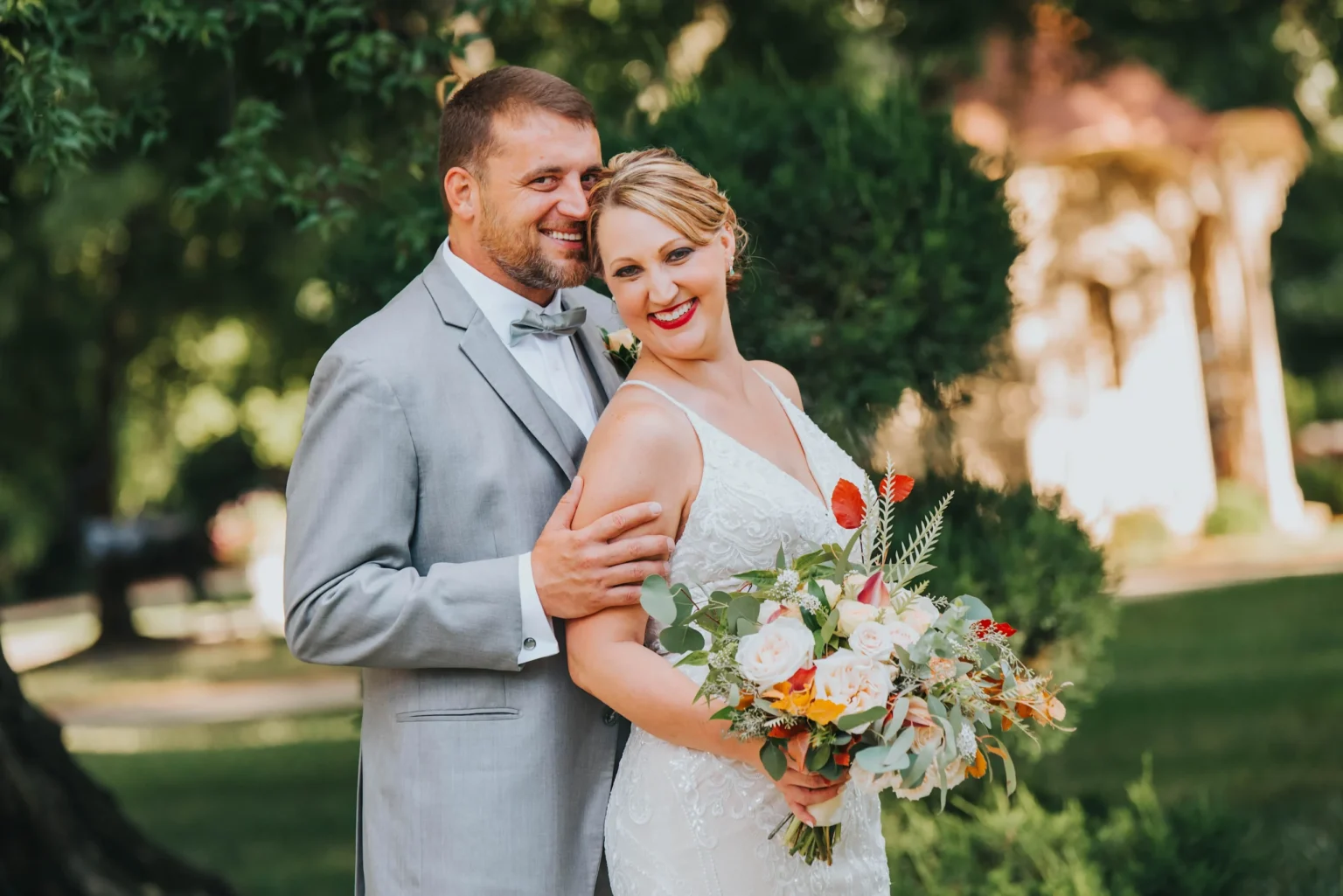 Elopement Wedding A couple stands closely together outdoors, both smiling happily. The man is behind the woman, dressed in a light gray suit with a bow tie. The woman is wearing a white wedding dress and holding a colorful bouquet of flowers. Lush greenery and trees fill the background. Elopements Inc