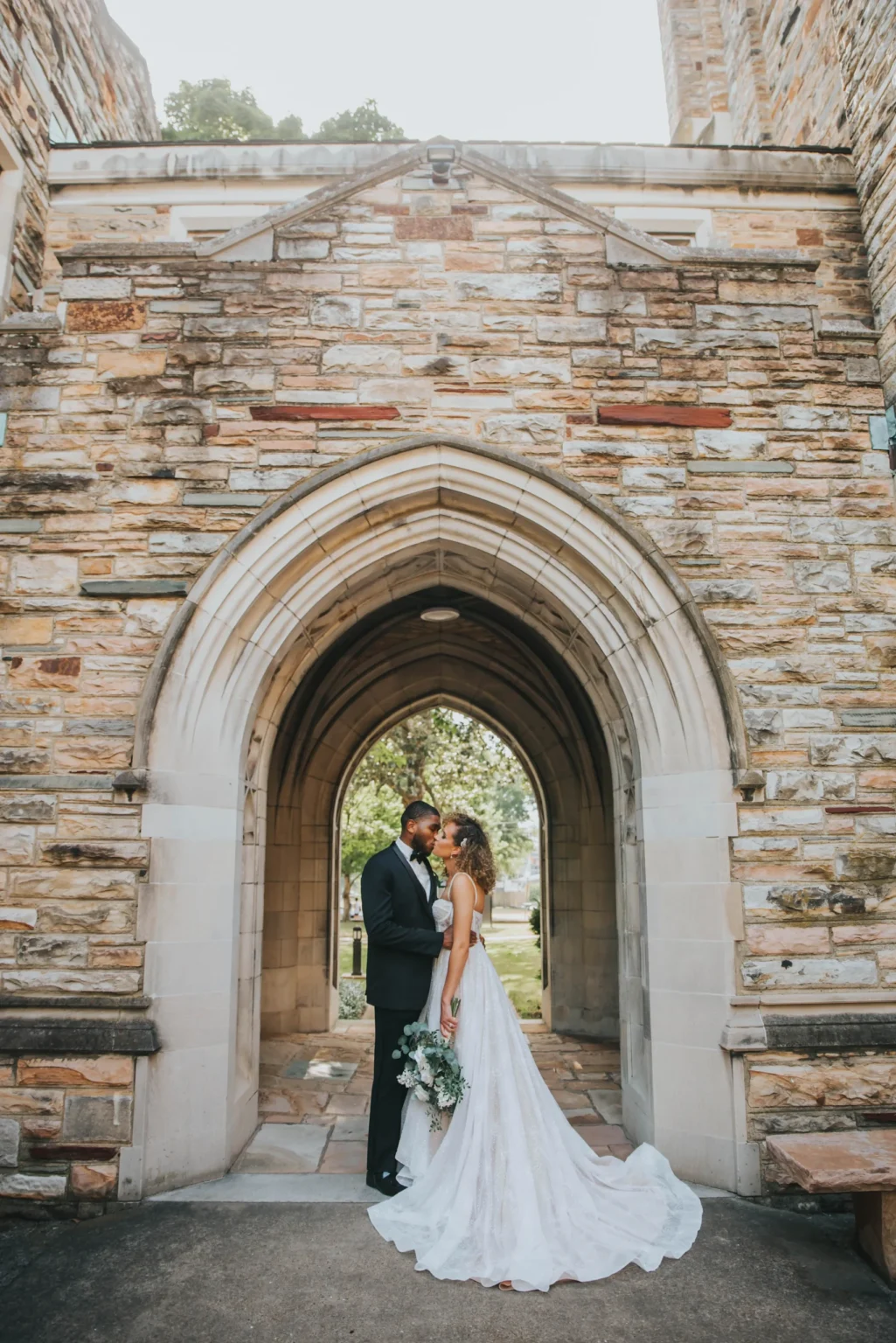 Elopement Wedding A wedding couple stands under a stone archway with detailed brickwork. The groom, in a black suit, and the bride, in a white gown holding a bouquet, share a tender moment with their foreheads touching. Sunlight filters through the archway, lighting up the scene and highlighting the bride’s train. Elopements Inc