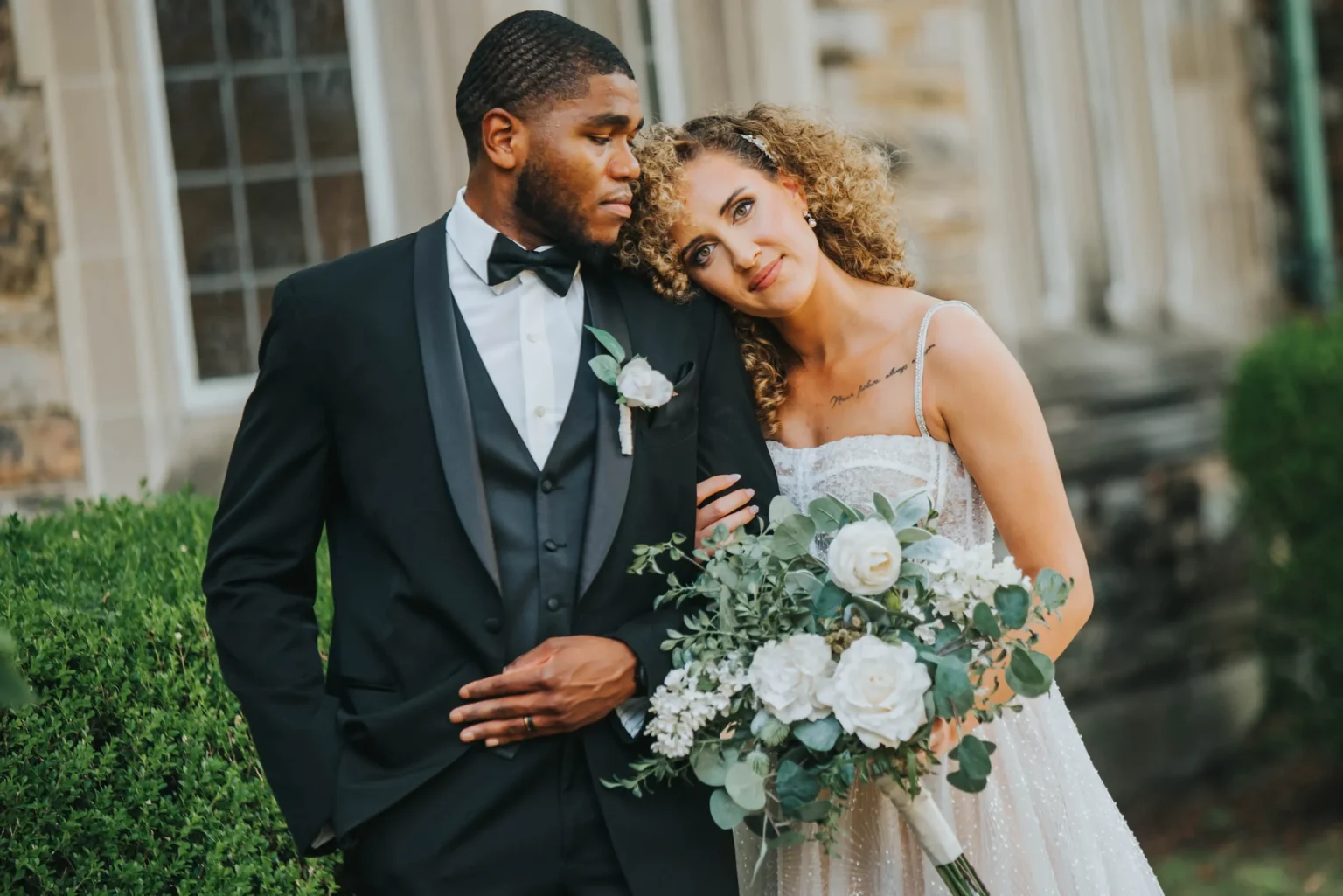 Elopement Wedding A couple dressed in formal wedding attire pose outdoors. The groom, in a black tuxedo with a bow tie, looks at the bride, who is in a white wedding gown and holding a bouquet of white flowers and greenery. They stand close together near a stone building with greenery in the background. Elopements Inc