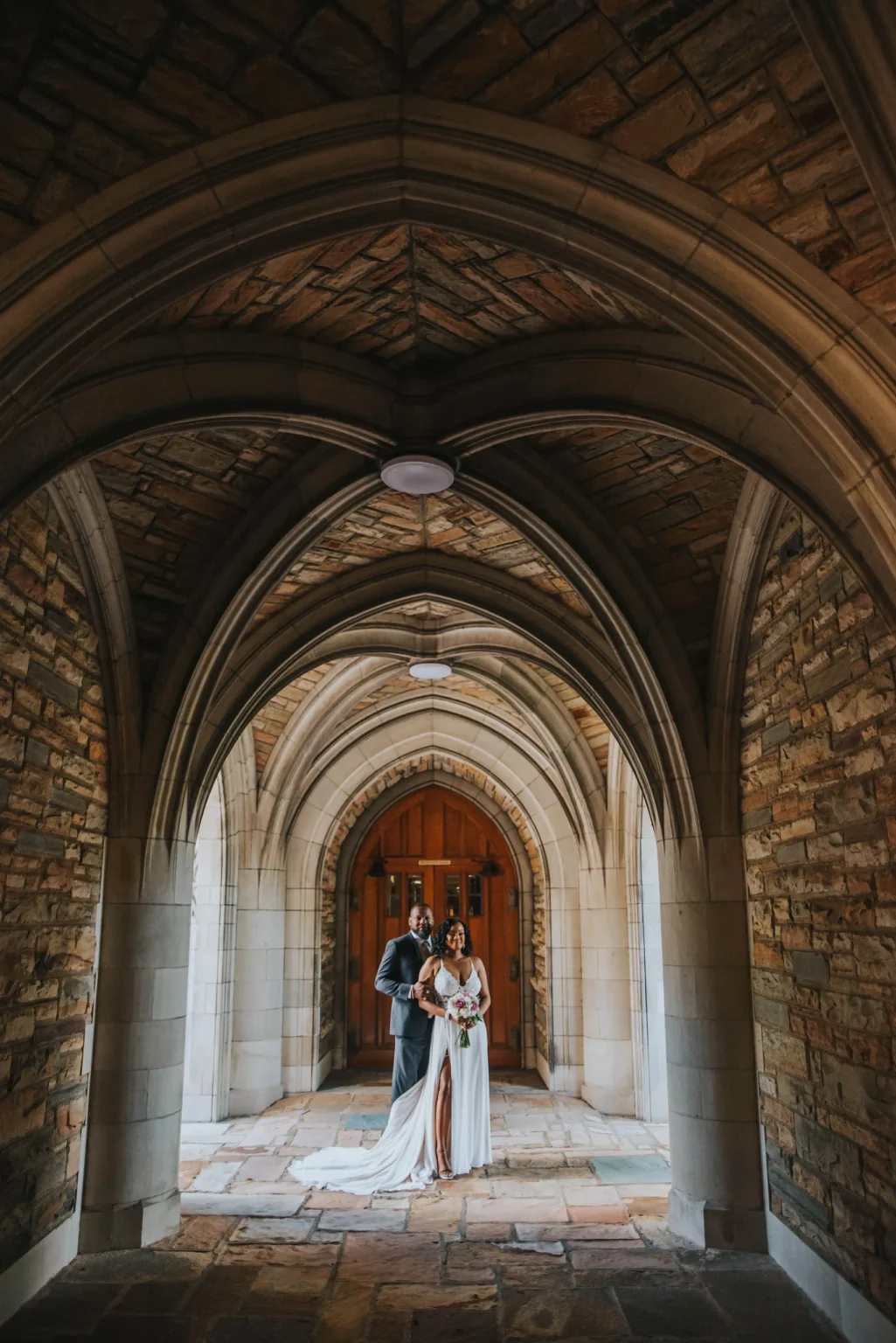 Elopement Wedding A couple stands under a stone archway in an embrace, dressed in wedding attire. The arched corridor features intricate stonework and a wooden door in the background. The bride holds a bouquet, and both smiles, creating a romantic and timeless scene. Elopements Inc