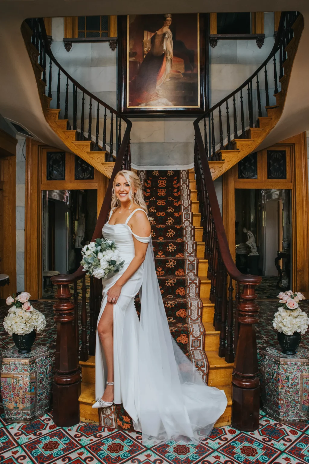 Elopement Wedding A bride in a white dress stands on a grand staircase, smiling, and holding a bouquet of white flowers. The staircase features wooden railings and patterned steps. Decorative vases with flowers are at the base. A large, formal portrait hangs above the landing, with intricate flooring visible below. Elopements Inc