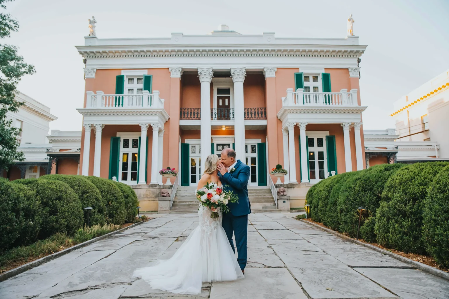 Elopement Wedding A newlywed couple stands in front of a grand, two-story mansion with large columns and a terracotta facade. The groom in a blue suit kisses the bride, who is in a white wedding dress and holding a colorful bouquet. They are standing on a stone walkway, flanked by manicured bushes. Elopements Inc