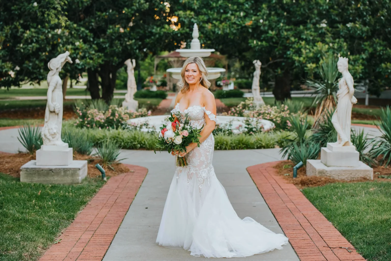 Elopement Wedding A bride in a strapless, white lace wedding gown stands smiling in front of a garden fountain. She holds a large bouquet of flowers with greenery. The garden features statues and lush foliage. The brick pathway beneath the bride leads to the ornate fountain surrounded by flowers. Elopements Inc