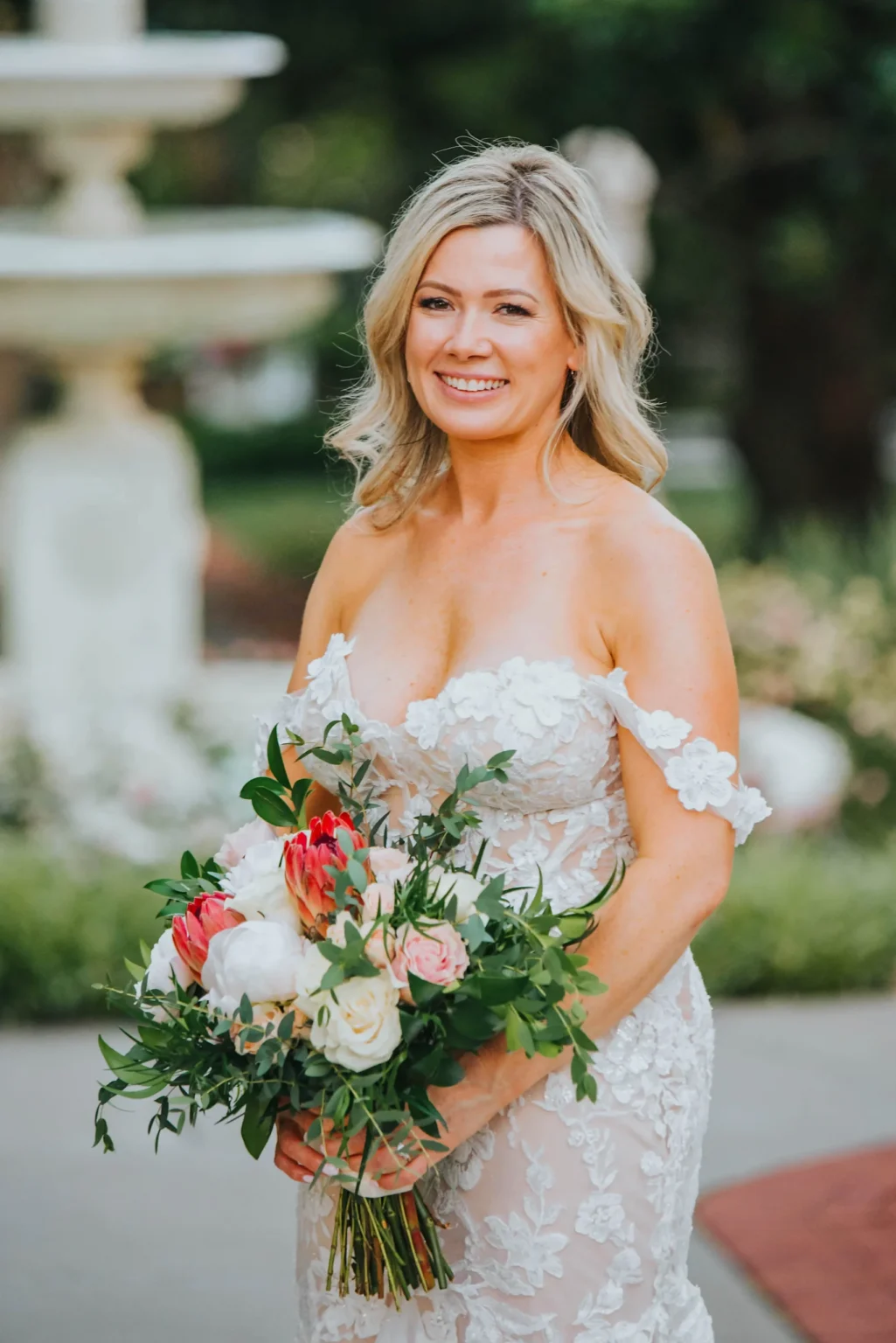 Elopement Wedding A bride stands outdoors in a garden, holding a lush bouquet of mixed flowers, including white and pink blooms with green foliage. She is wearing an off-the-shoulder, floral lace wedding dress with a sweetheart neckline. Her blonde hair is styled in soft waves, and she is smiling gently at the camera. Elopements Inc