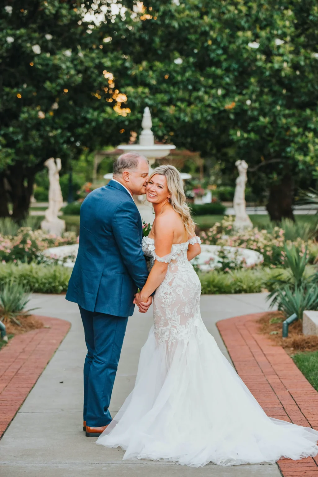 Elopement Wedding A bride and groom stand hand in hand on a garden path with greenery and statues in the background. The groom wears a blue suit and the bride wears an elegant, white off-the-shoulder gown with lace details. Both are smiling, with the bride looking at the camera while the groom leans in close. Elopements Inc