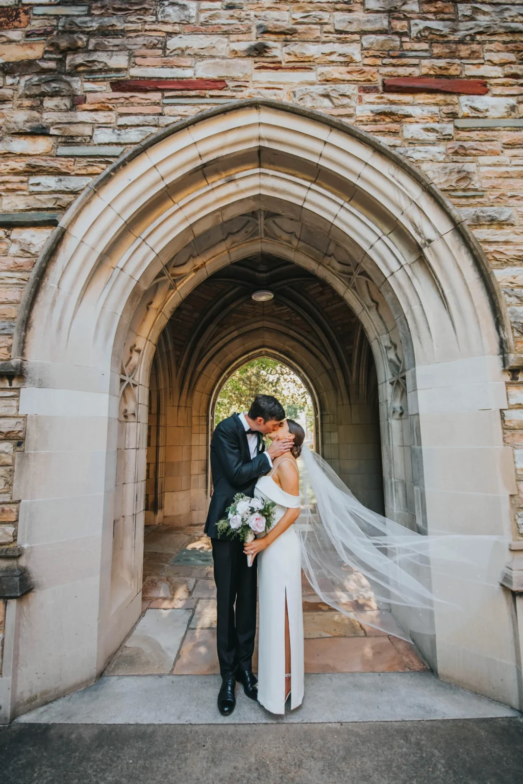 Elopement Wedding A bride and groom share a kiss under a grand stone archway. The bride wears a white gown with her veil flowing to the side, and holds a bouquet of flowers. The groom is in a black suit and white dress shirt. The scene exudes romance and elegance against the backdrop of the textured stone architecture. Elopements Inc