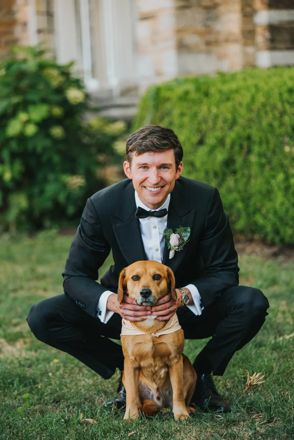 Elopement Wedding A man dressed in a black tuxedo with a white shirt and black bow tie is squatting down on a lawn, smiling, with his hands around a brown dog wearing a matching bow tie. The background features greenery and a blurred section of a stone building. Elopements Inc