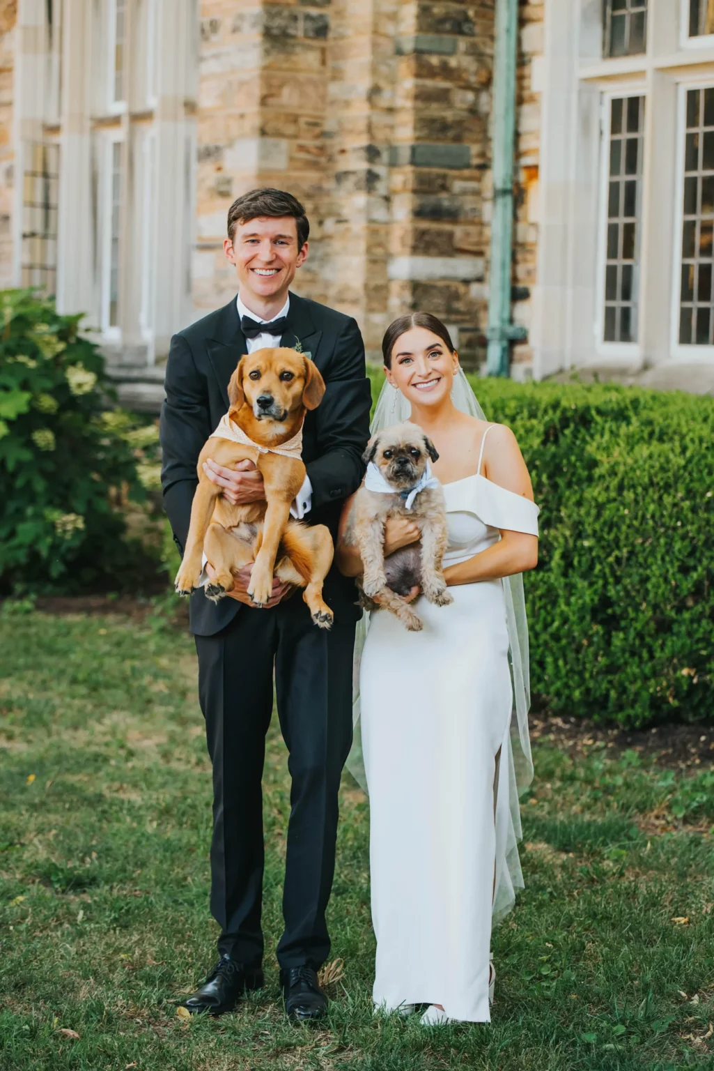 Elopement Wedding A couple, dressed in formal wedding attire, stands outdoors in front of a stone building with large windows. The man, in a black tuxedo, holds a brown dog. The woman, in a white off-shoulder gown, holds a small grey dog. Both are smiling. Green bushes and lawn surround them. Elopements Inc