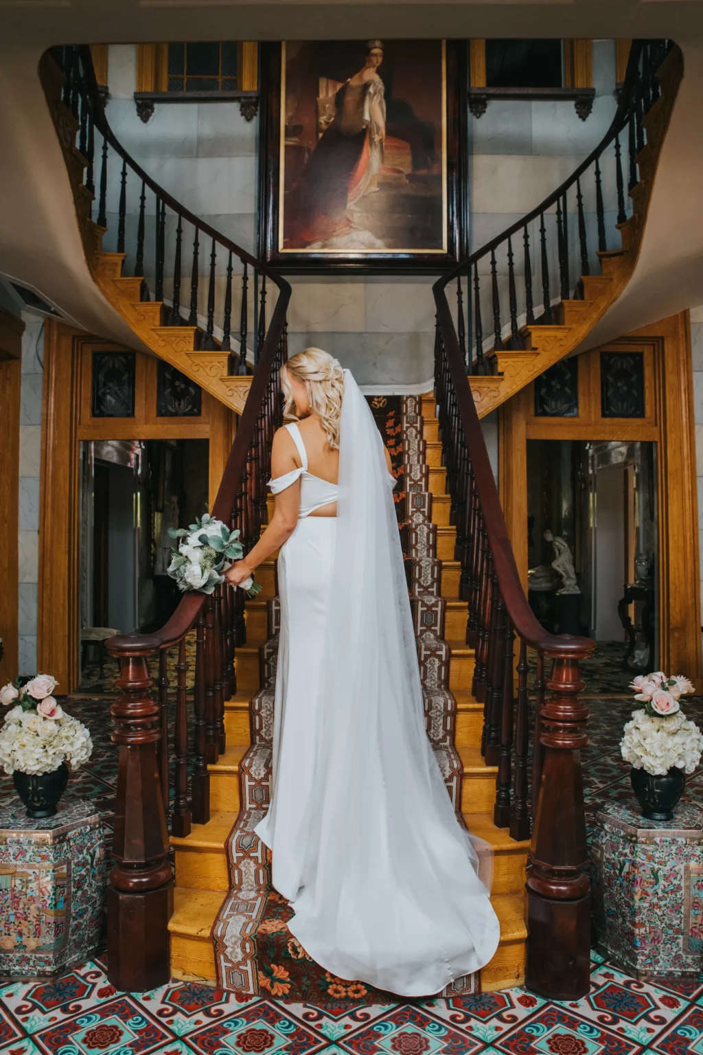 Elopement Wedding A bride, wearing a flowing white dress and veil, stands at the base of a grand staircase with polished wooden railings. She holds a bouquet of flowers and looks up, her back to the camera. The floor features intricate patterned tiles, and a large painting hangs at the top of the stairs. Elopements Inc