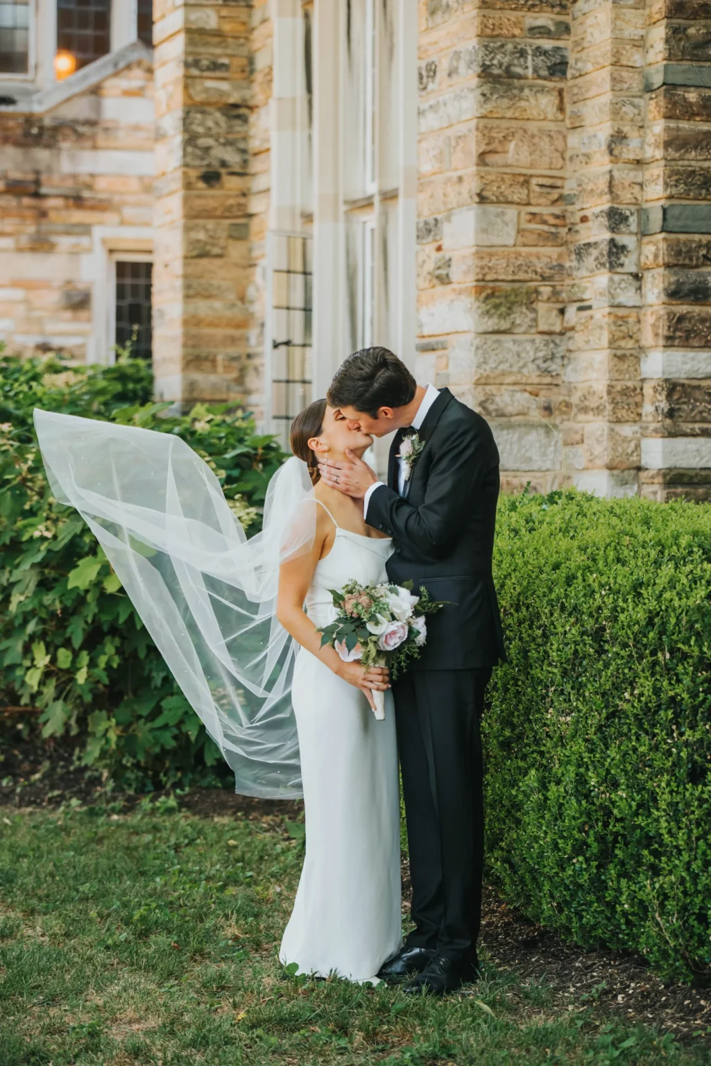 Elopement Wedding A couple, dressed in wedding attire, shares a kiss in front of a stone building. The groom, in a black suit, holds the bride, who is in a white dress with a veil flowing behind her. The bride holds a bouquet of flowers. Lush greenery and bushes are in the background. Elopements Inc