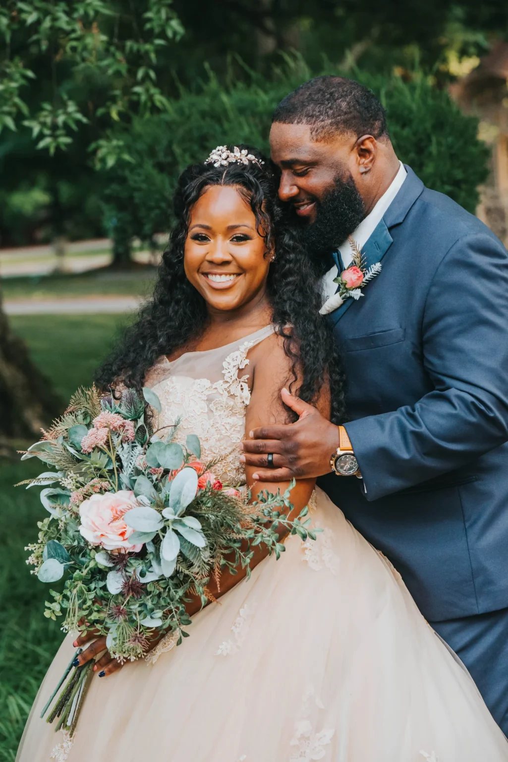 Elopement Wedding A couple on their wedding day stands outdoors, smiling radiantly. The bride in a lace-embellished gown holds a lush bouquet of flowers, while the groom, dressed in a dark suit, lovingly embraces her from behind. Trees and greenery are visible in the background, adding to the romantic atmosphere. Elopements Inc