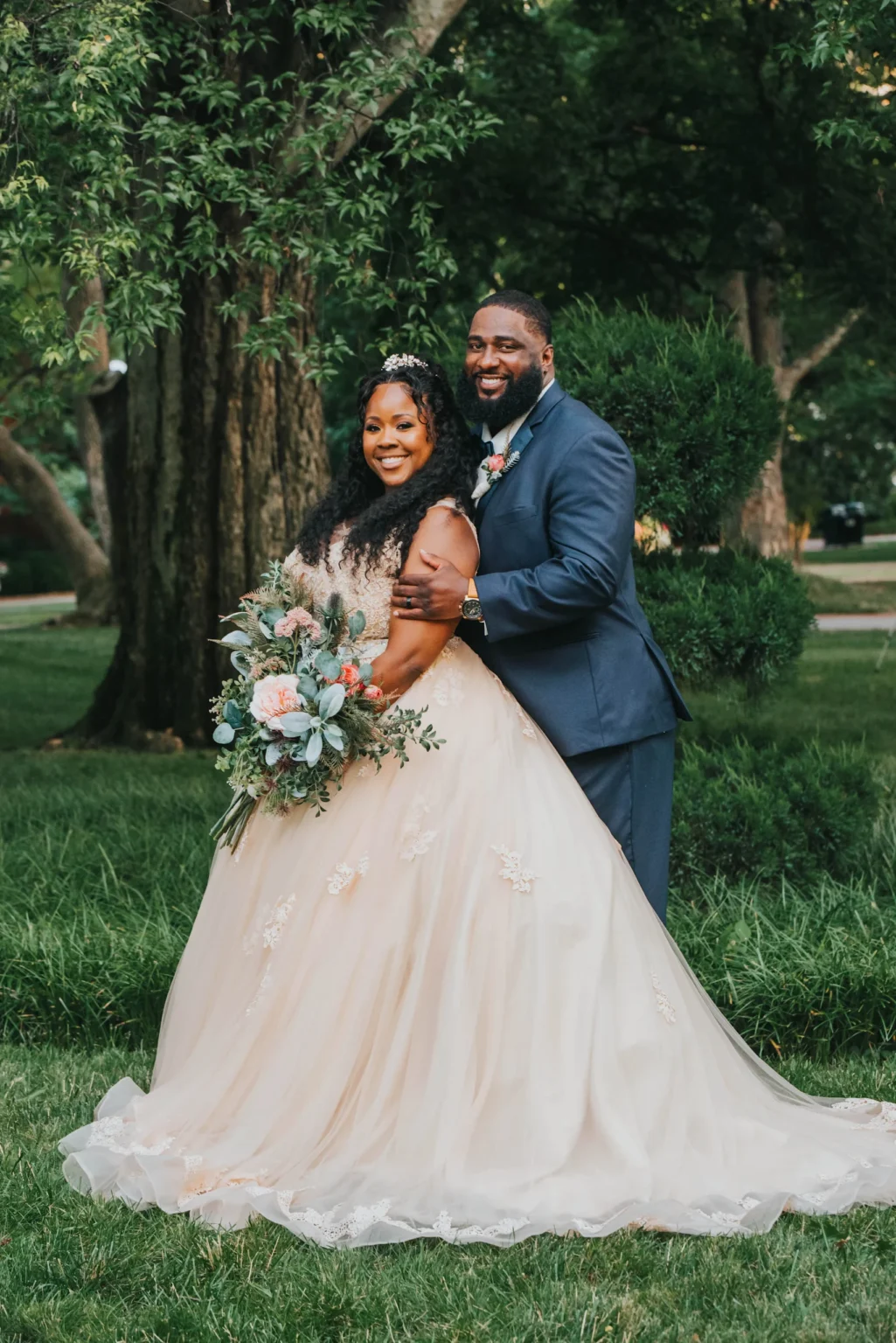 Elopement Wedding A smiling bride in an ivory, floral-embellished gown holds a bouquet of pink and white flowers, standing beside the groom in a navy blue suit. They are outdoors in a lush, green garden with trees in the background, embracing each other happily. Elopements Inc