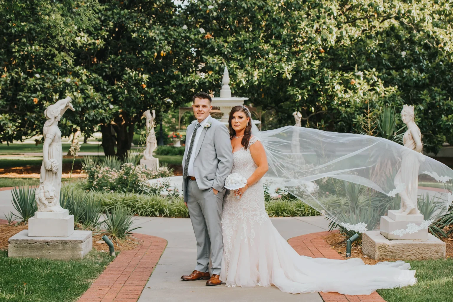 Elopement Wedding A bride and groom stand together in a garden for a wedding photo. The groom wears a light gray suit and brown shoes, while the bride wears a flowing white lace dress with a long veil that is gently blowing in the wind. They are surrounded by greenery, statues, and a white stone pathway. Elopements Inc