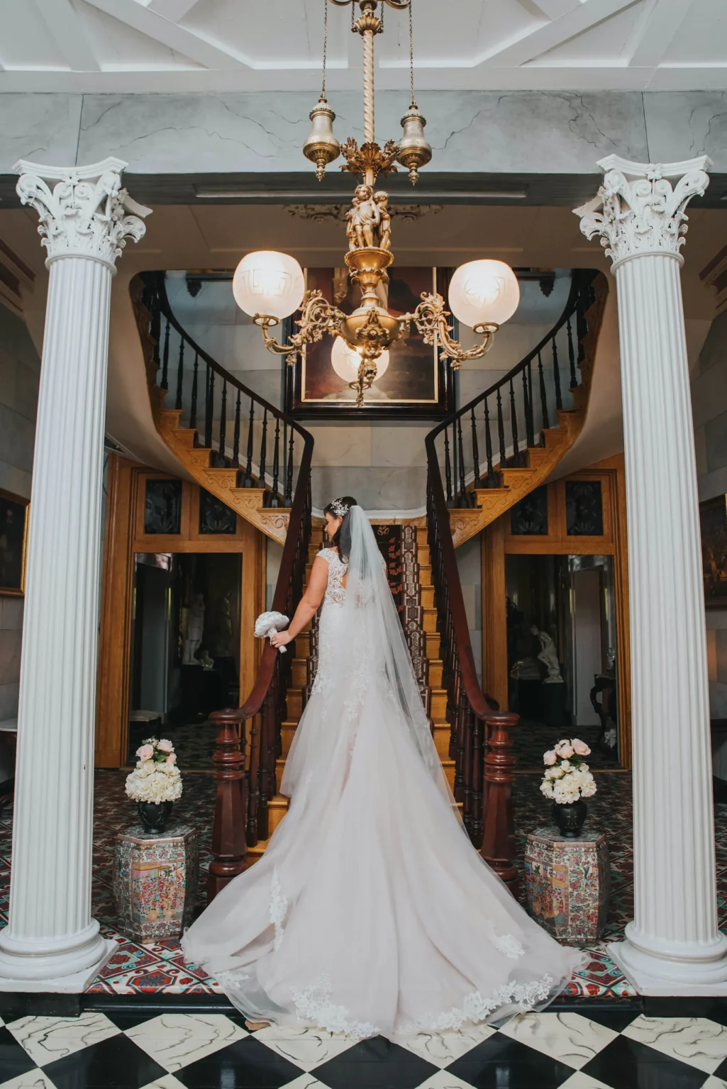 Elopement Wedding A bride in a white gown with a long train stands at the foot of a grand wooden staircase adorned with floral arrangements. She faces away from the camera, holding a bouquet, under a chandelier hanging from a high ceiling with ornate columns framing the stairs. Elopements Inc