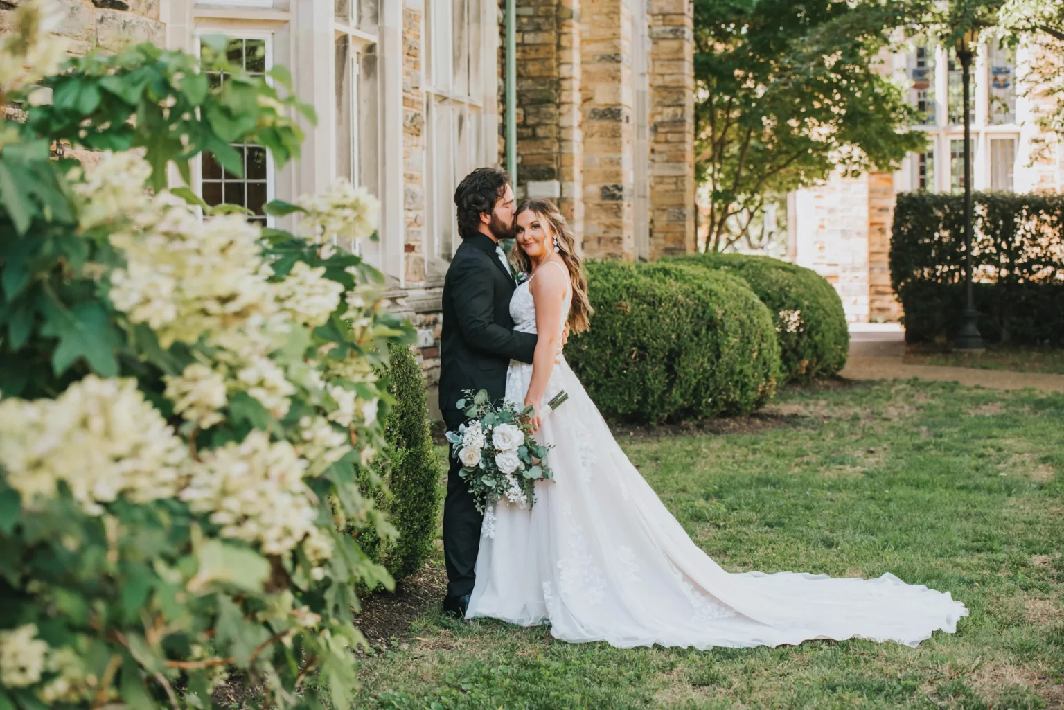 Elopement Wedding A couple dressed in wedding attire stands close together outside an old stone building with arched windows. The bride holds a bouquet of white and blush flowers and wears a flowing white gown. The groom, in a dark suit, gently kisses her forehead. Green foliage surrounds them. Elopements Inc