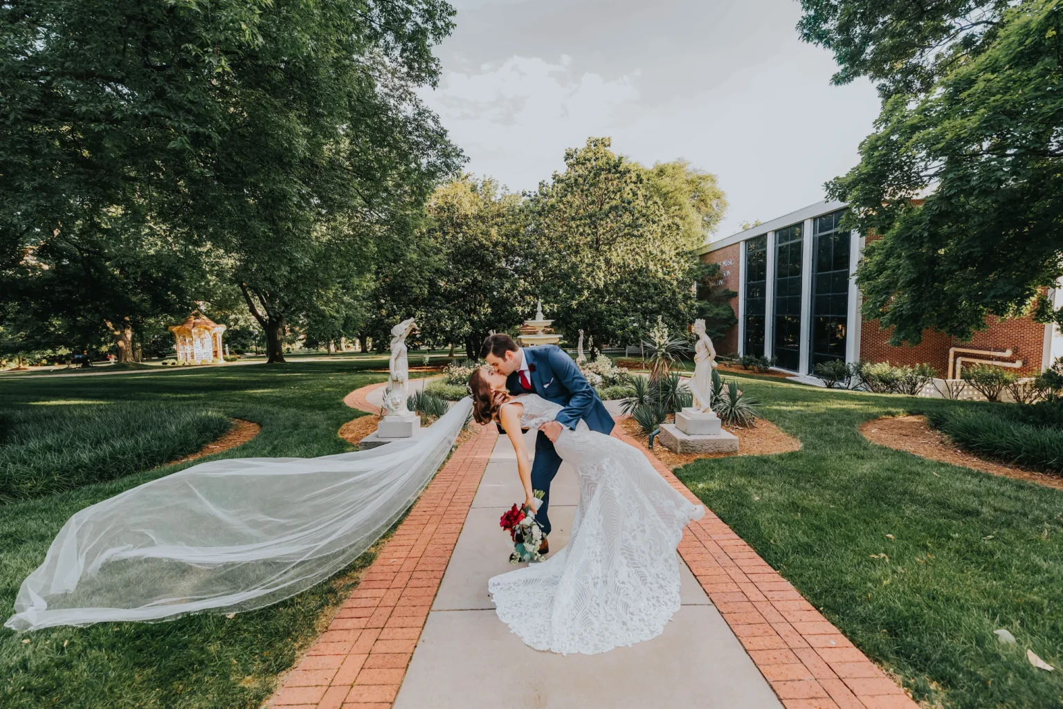 Elopement Wedding A groom in a blue suit and bride in a white, lacy dress share a kiss as he dips her back on a garden pathway. The bride's long veil cascades behind, and she holds a bouquet of red flowers. They are surrounded by greenery, statues, and a brick building is visible in the background. Elopements Inc