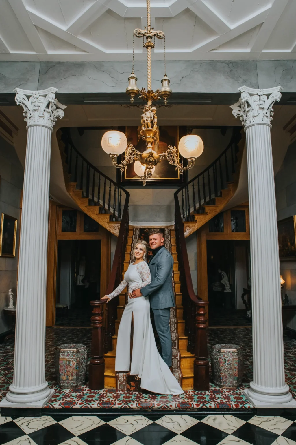 Elopement Wedding A couple stands close together on a staircase, posing for a formal photo. The woman is in a long white dress, and the man is in a gray suit. They are flanked by two elegant columns with a chandelier overhead, set against a backdrop of classic architectural details and patterned flooring. Elopements Inc