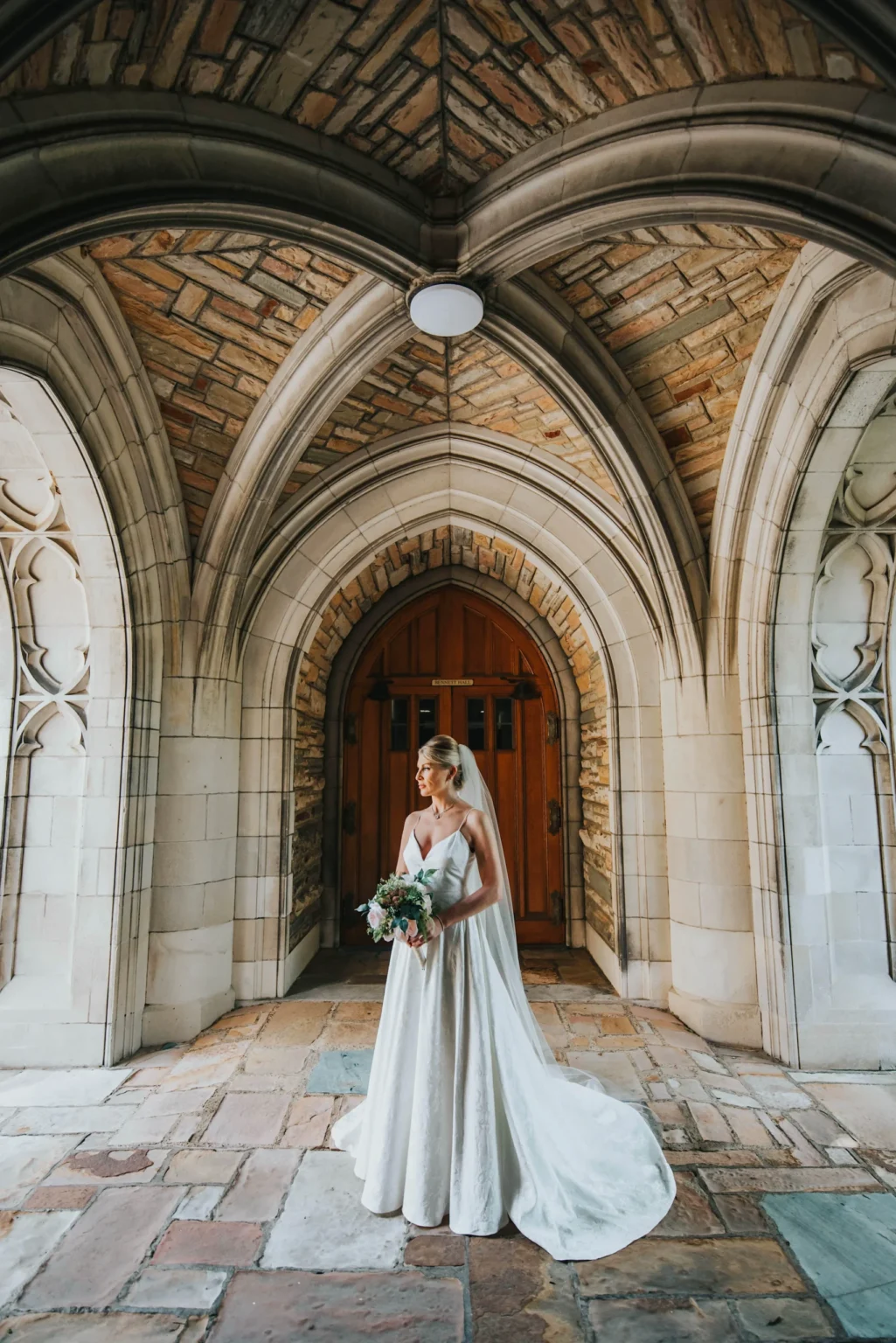 Elopement Wedding A bride in a white wedding dress stands under a stone archway. She holds a bouquet of flowers and gazes to her left. The archway features intricate stonework, with pointed arches and detailed carvings, creating a historic and elegant backdrop. The floor is paved with large stones. Elopements Inc