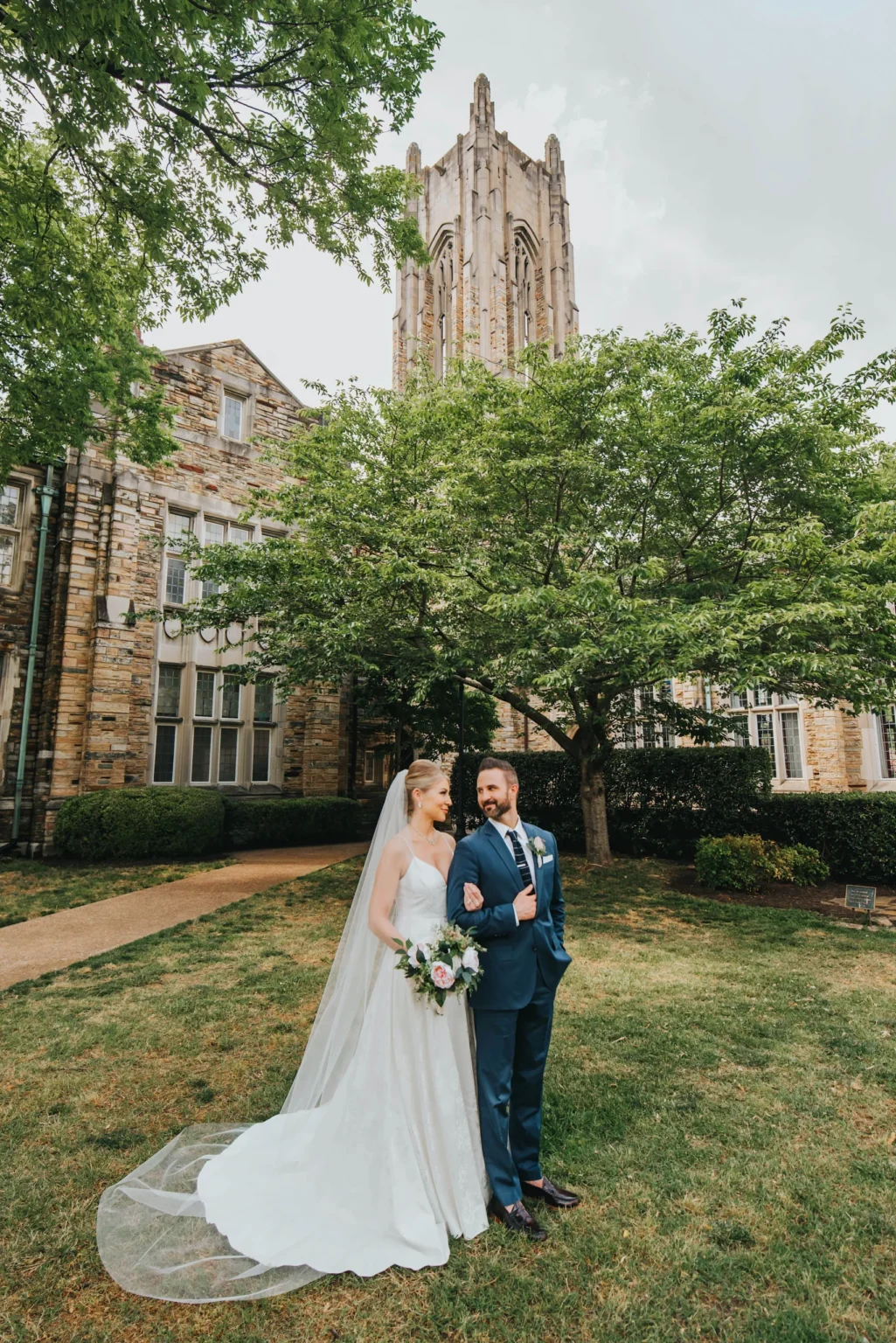 Elopement Wedding A newlywed couple stands arm-in-arm outdoors, smiling at each other. The bride wears a white gown with a long veil and holds a bouquet of flowers. The groom is in a blue suit with a bow tie. They are standing on a manicured lawn in front of a stone building with large windows and a tall tower. Elopements Inc