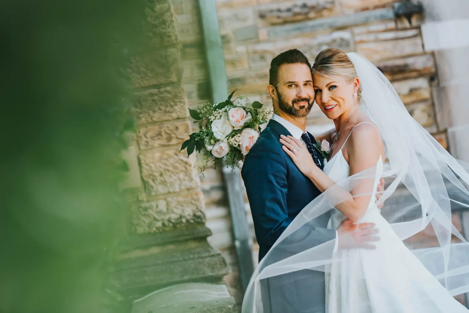 Elopement Wedding A smiling bride and groom stand close to each other in front of a stone building. The bride, wearing a white dress and veil, holds a bouquet. The groom is dressed in a dark suit. Both look happily at the camera, with the bride's veil flowing and greenery slightly blurred in the foreground. Elopements Inc