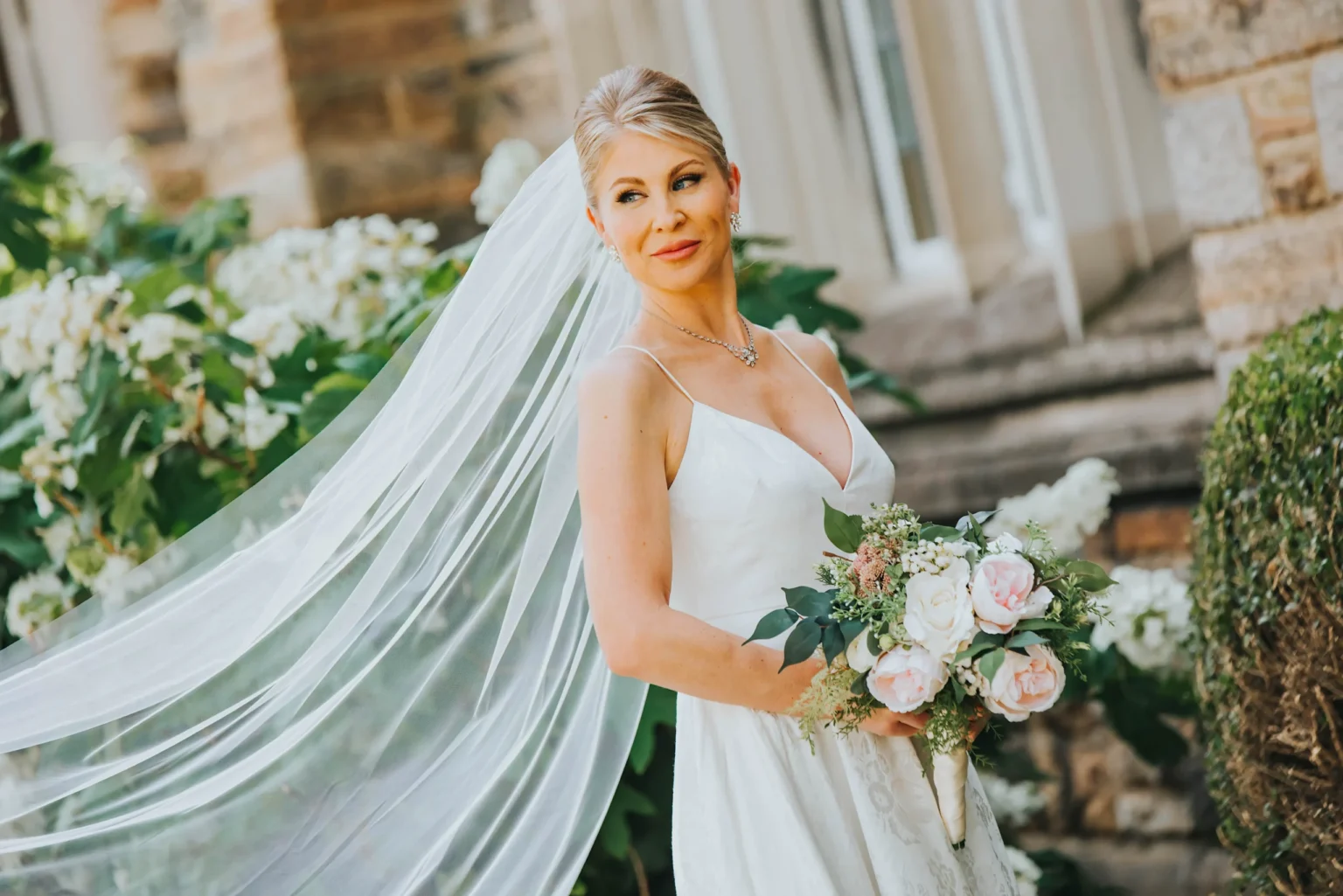 Elopement Wedding A bride stands outside, wearing a sleeveless white wedding dress with a long, flowing veil. She holds a bouquet of pink and white flowers and smiles softly. Behind her are green bushes and a stone building, giving a serene and romantic atmosphere. Elopements Inc